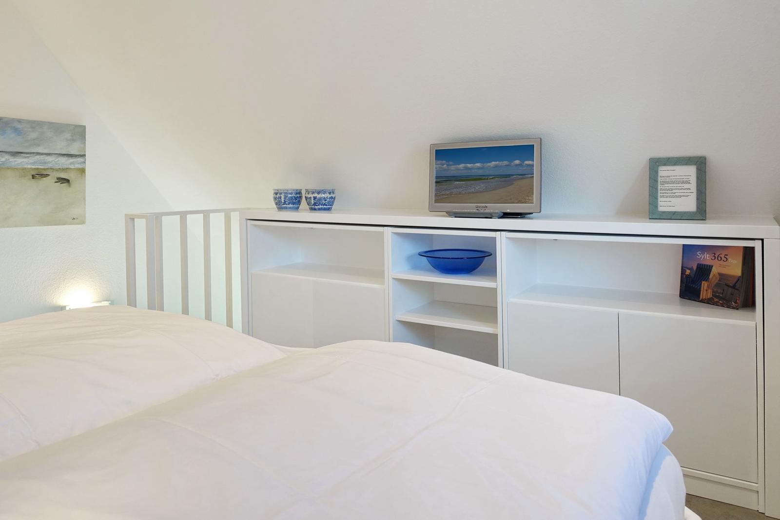Bedroom with white bed and shelf. On the shelf are two bowls, a TV, and a picture frame.