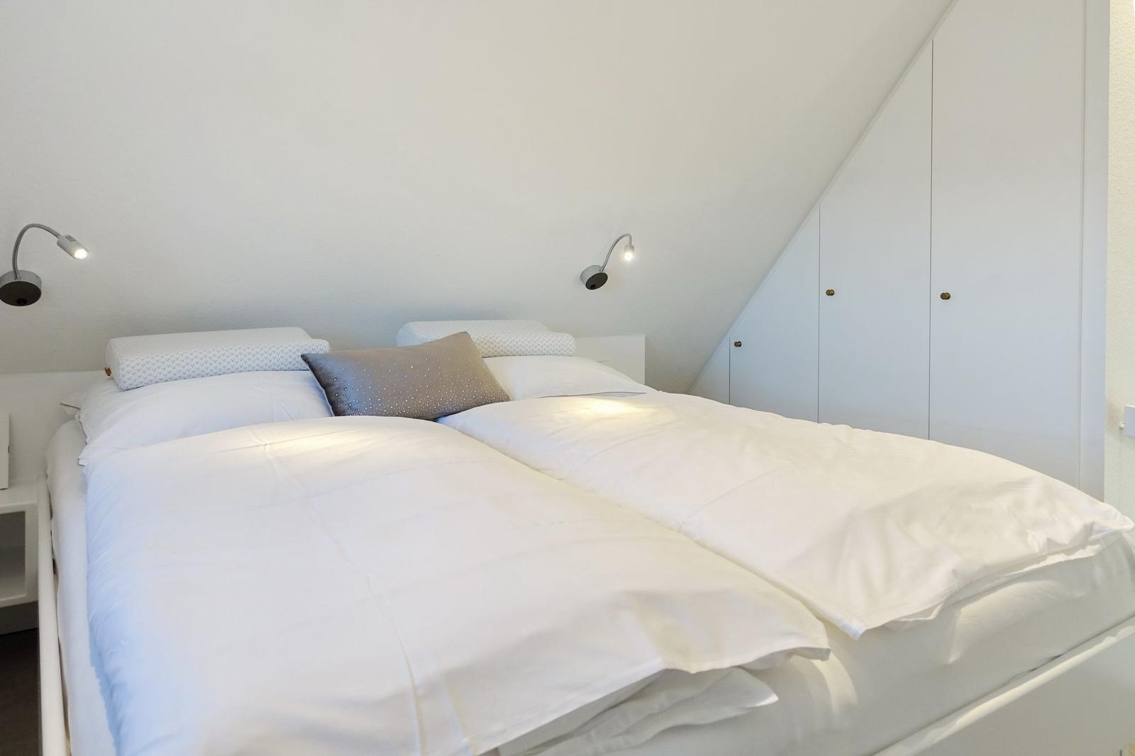 Bedroom with double bed, white bedding, and two wall lamps. A white wardrobe is against the wall.