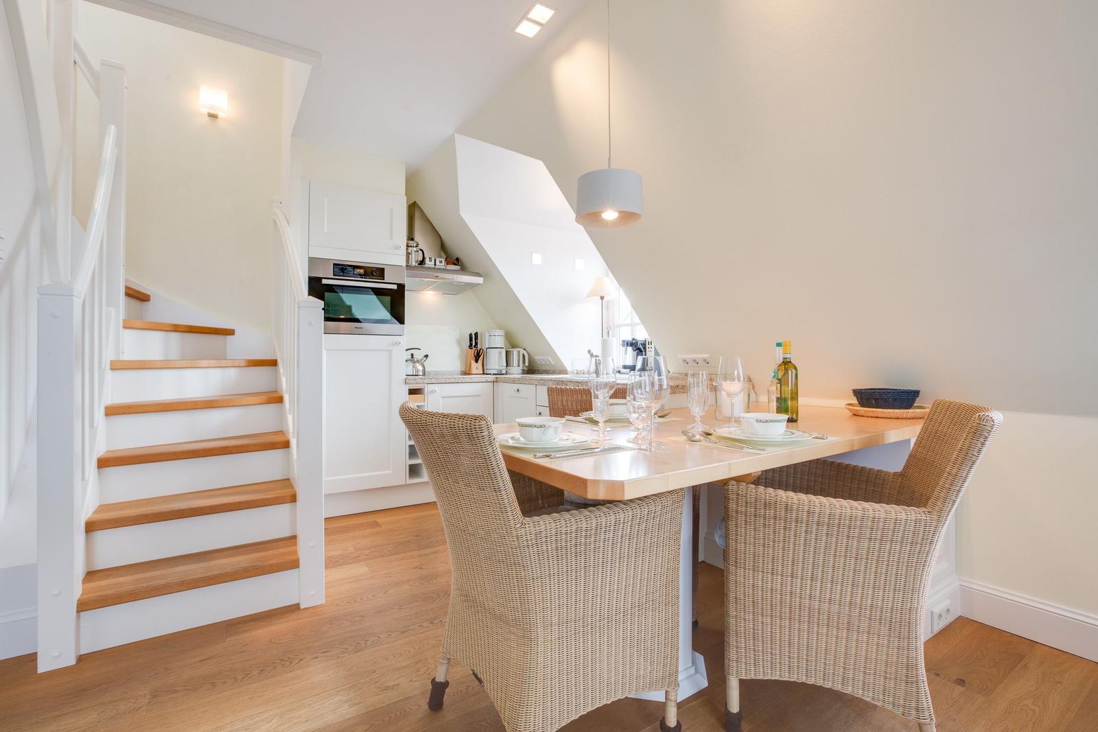 Dining area with table and chairs, kitchen, and staircase in background.