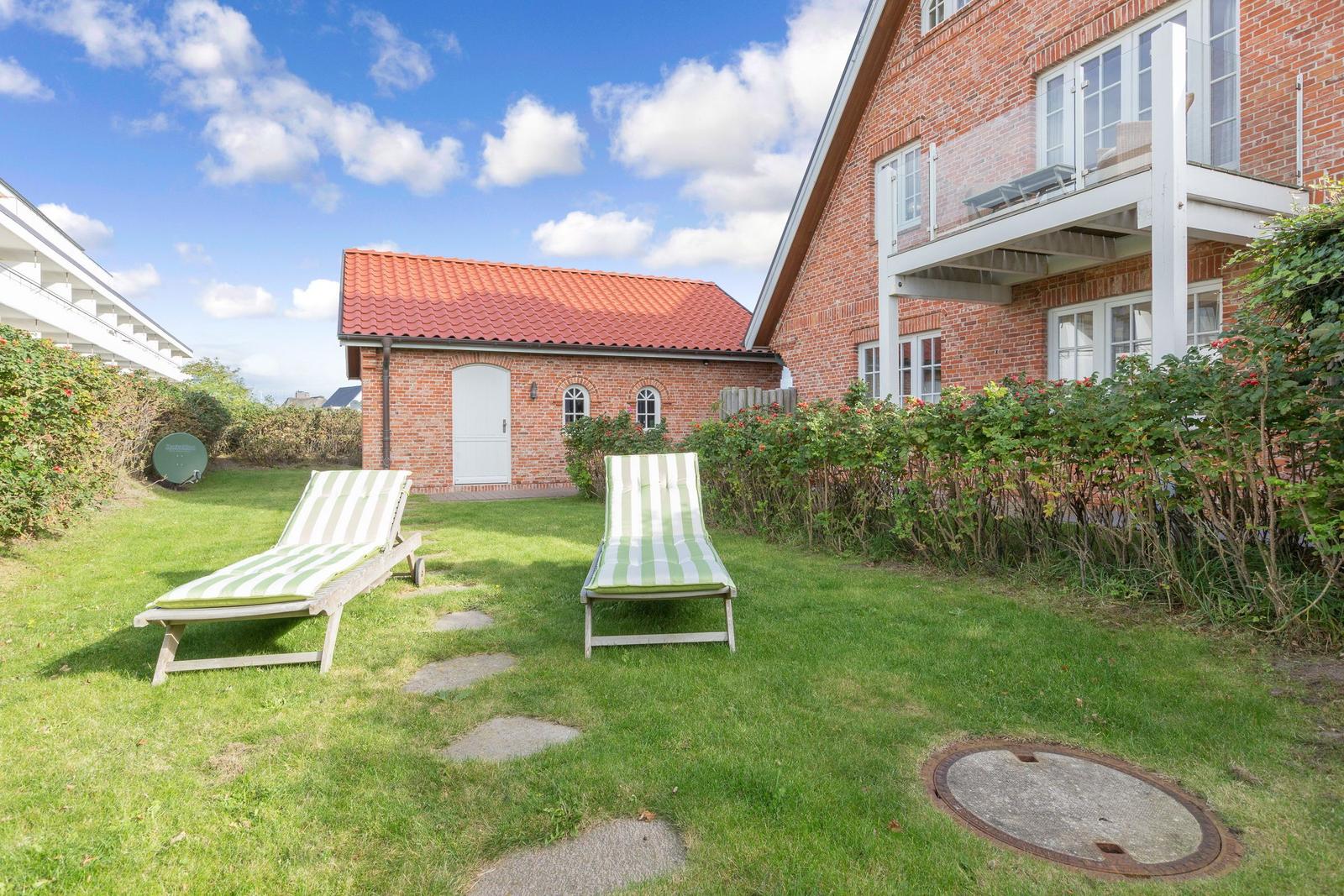 Green garden with two lounge chairs in front of a brick house under blue sky.