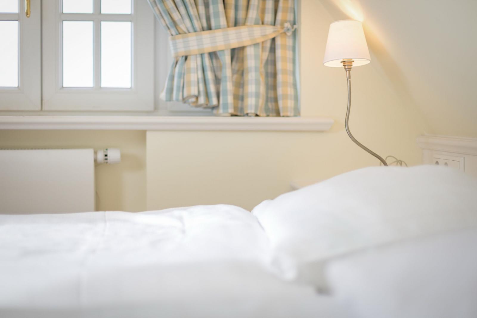 Bed with white linens, wall lamp, and window with checkered curtains.