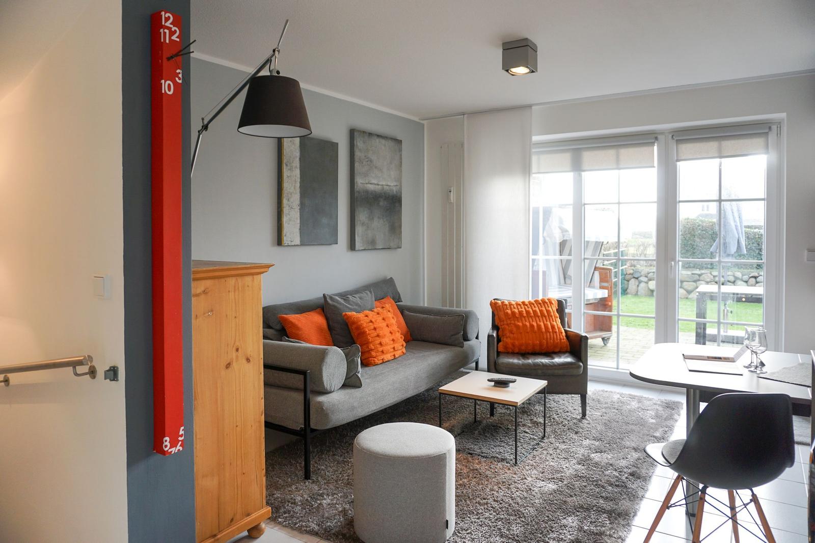 Living room with sofa, armchair, and dining table. Red wall clock and large windows.