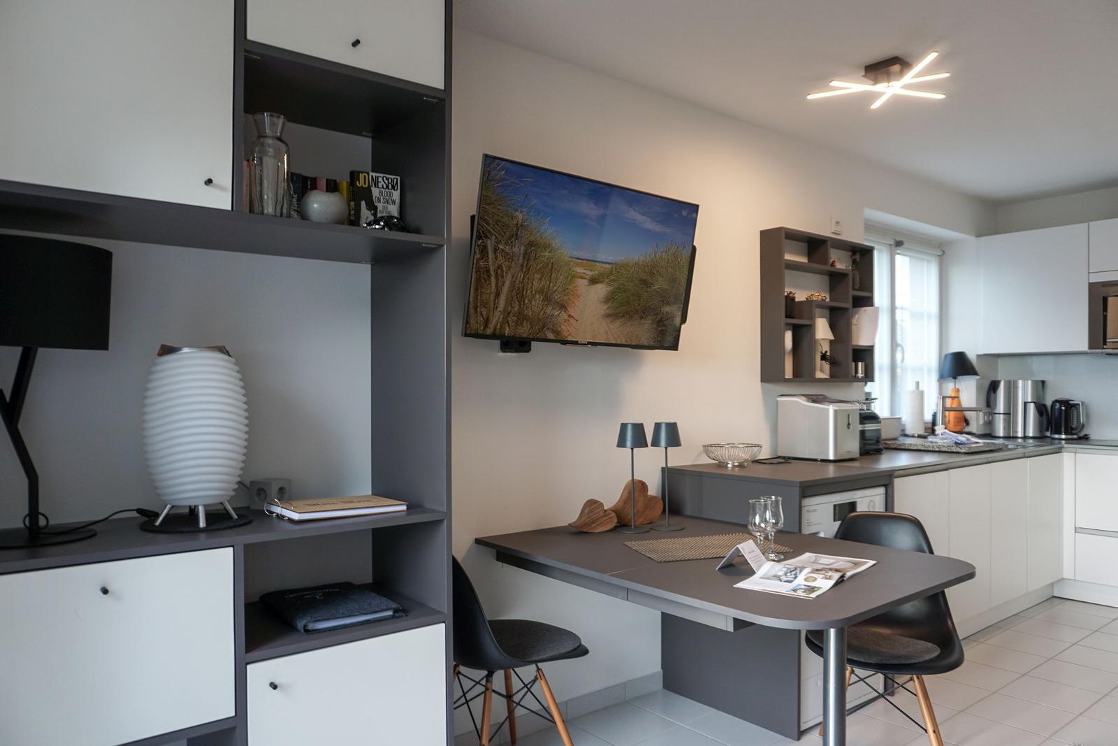 Kitchen with dining table, TV, and shelves. Modern furnishings with white and gray furniture.