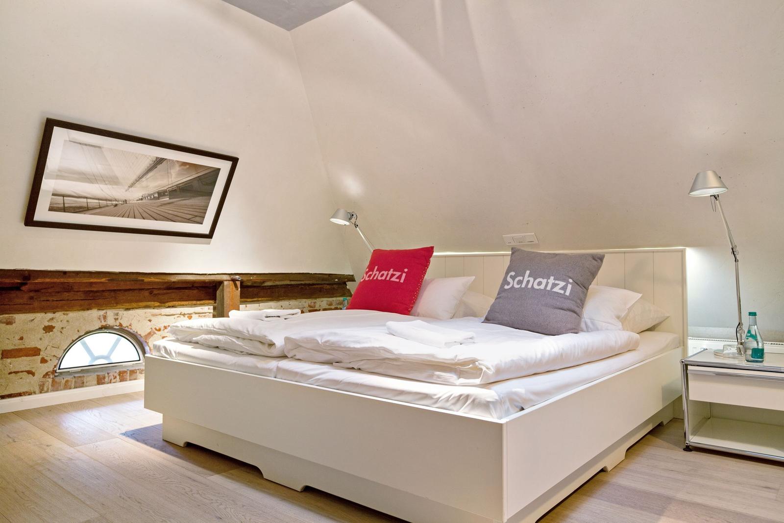Schlafzimmer mit weißem Bett, rotem und grauem Kissen, Nachttisch und Wandbild.