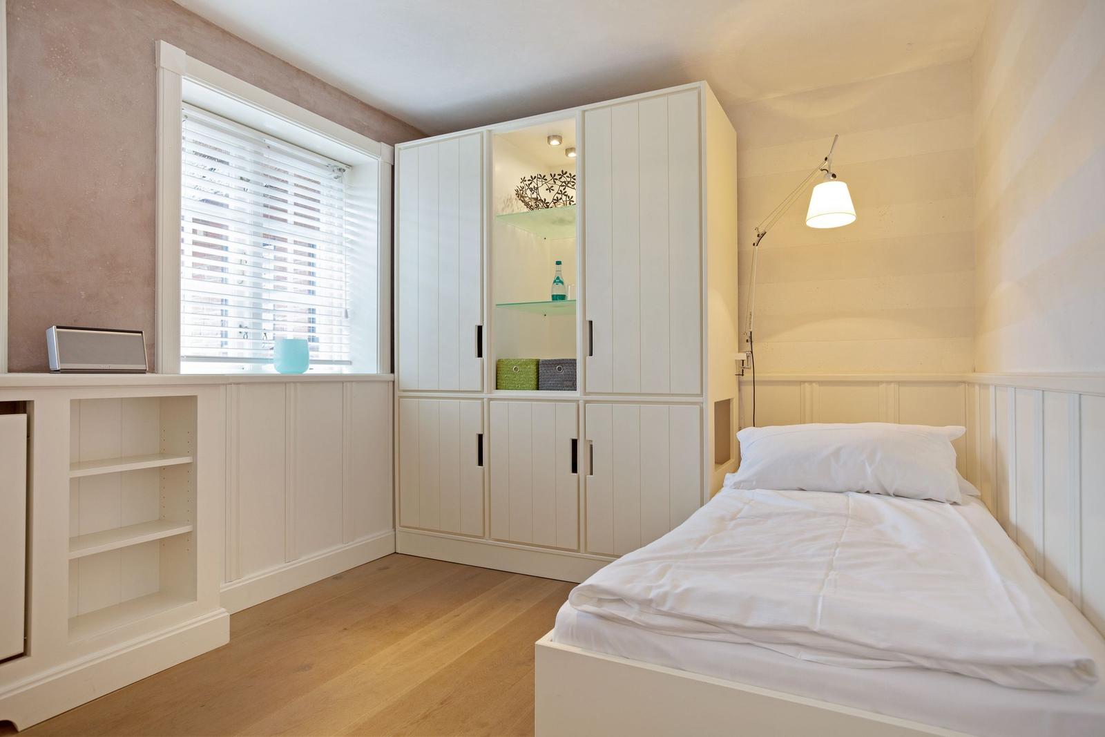 Schlafzimmer mit weißem Bett, Kleiderschrank und Fenster mit Jalousie.