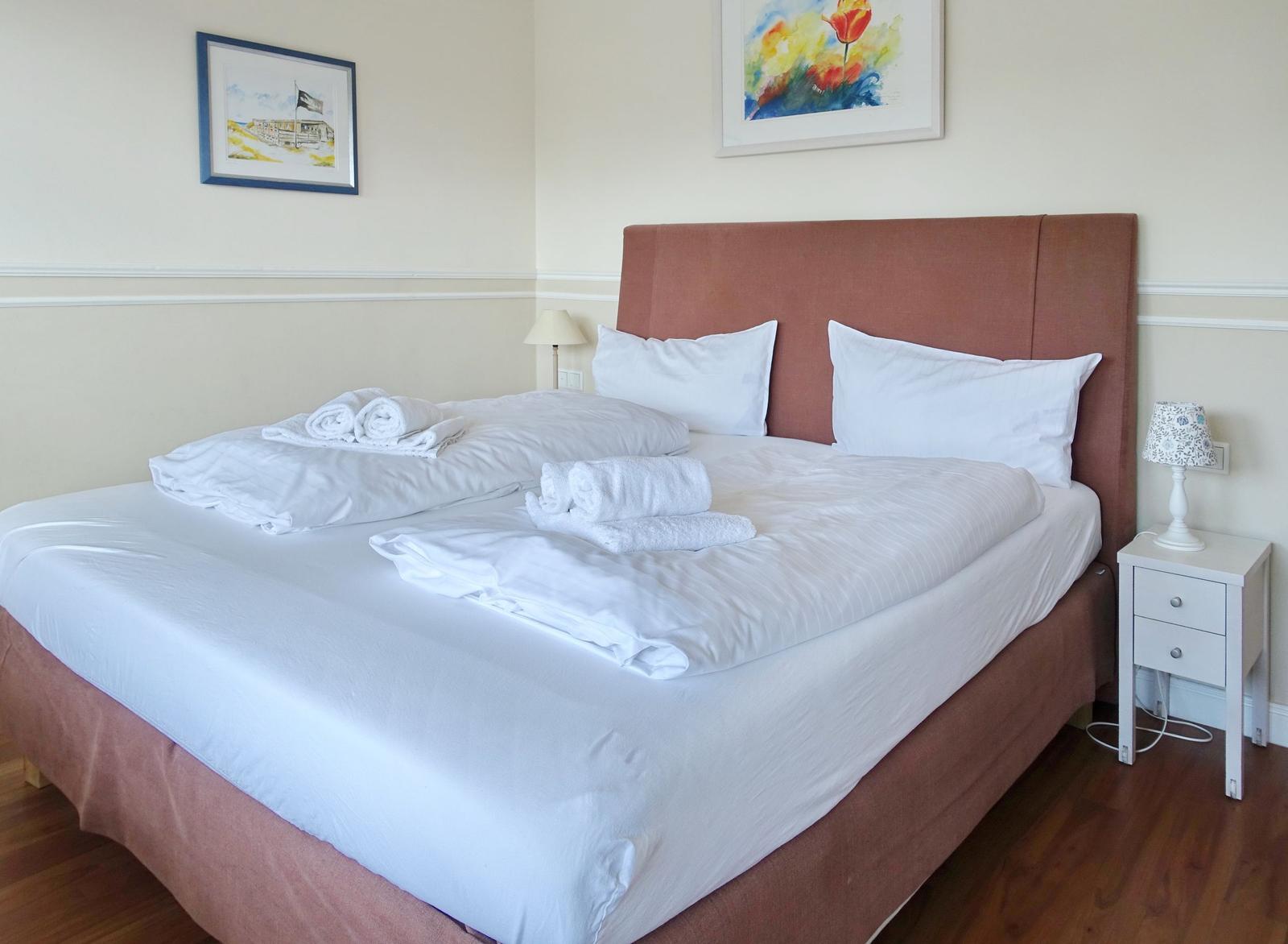 Ein Bett mit weißen Laken und Handtüchern. An der Wand hängen zwei Bilder.