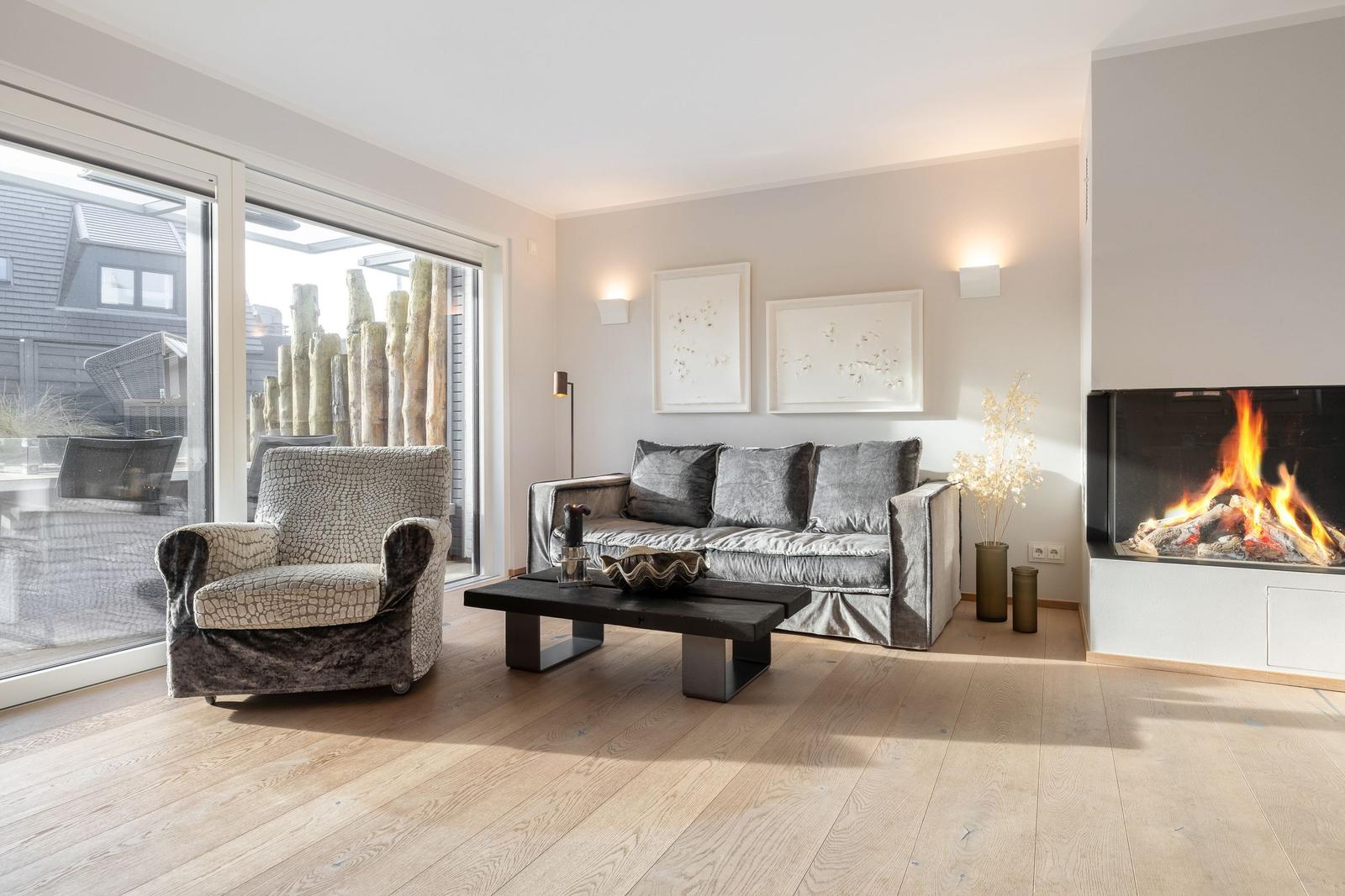 Living room with sofa, armchair, and fireplace. Wooden floor and large windows.