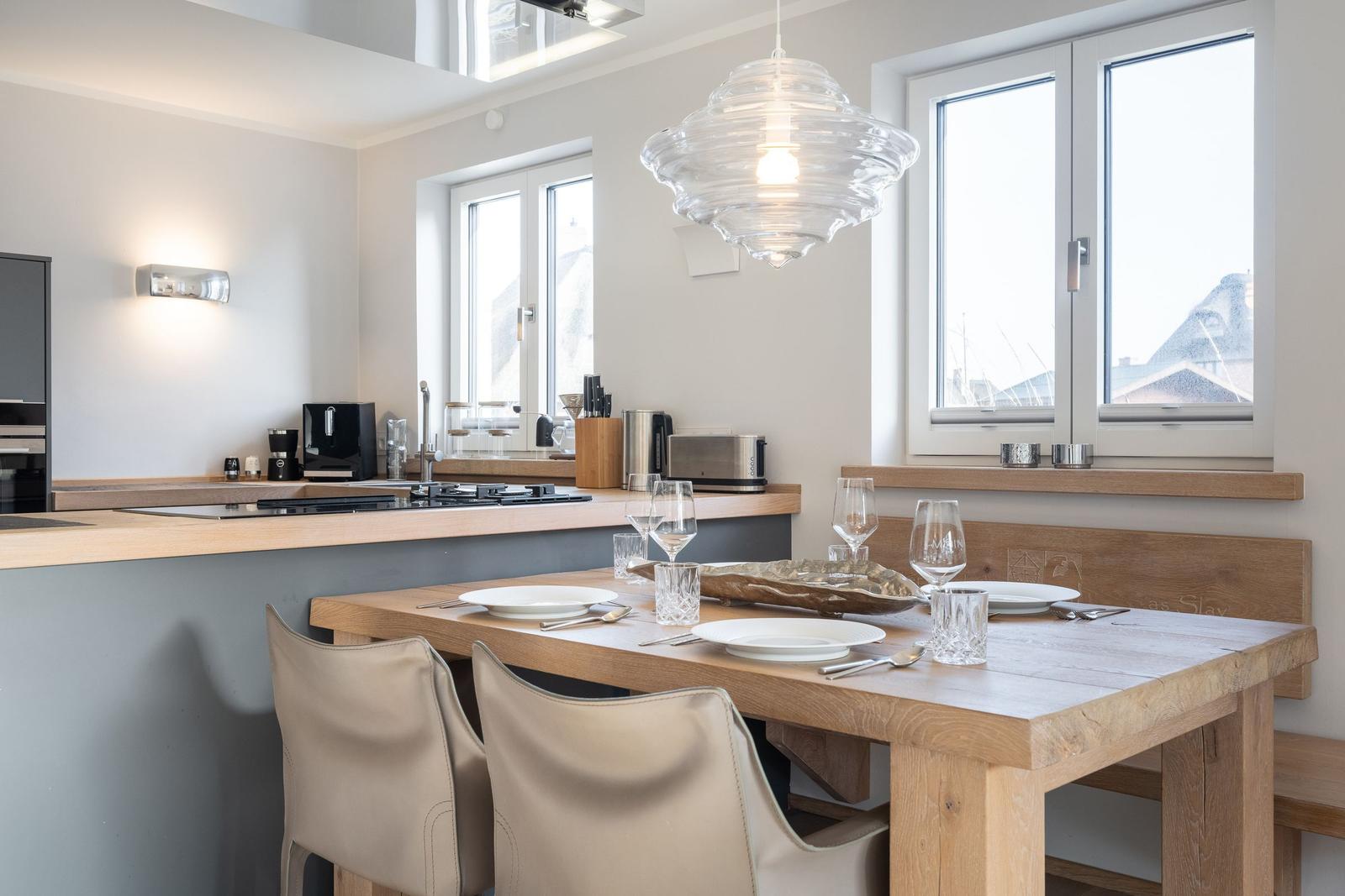 Dining area with wooden table, chairs, and windows. Kitchen in background.