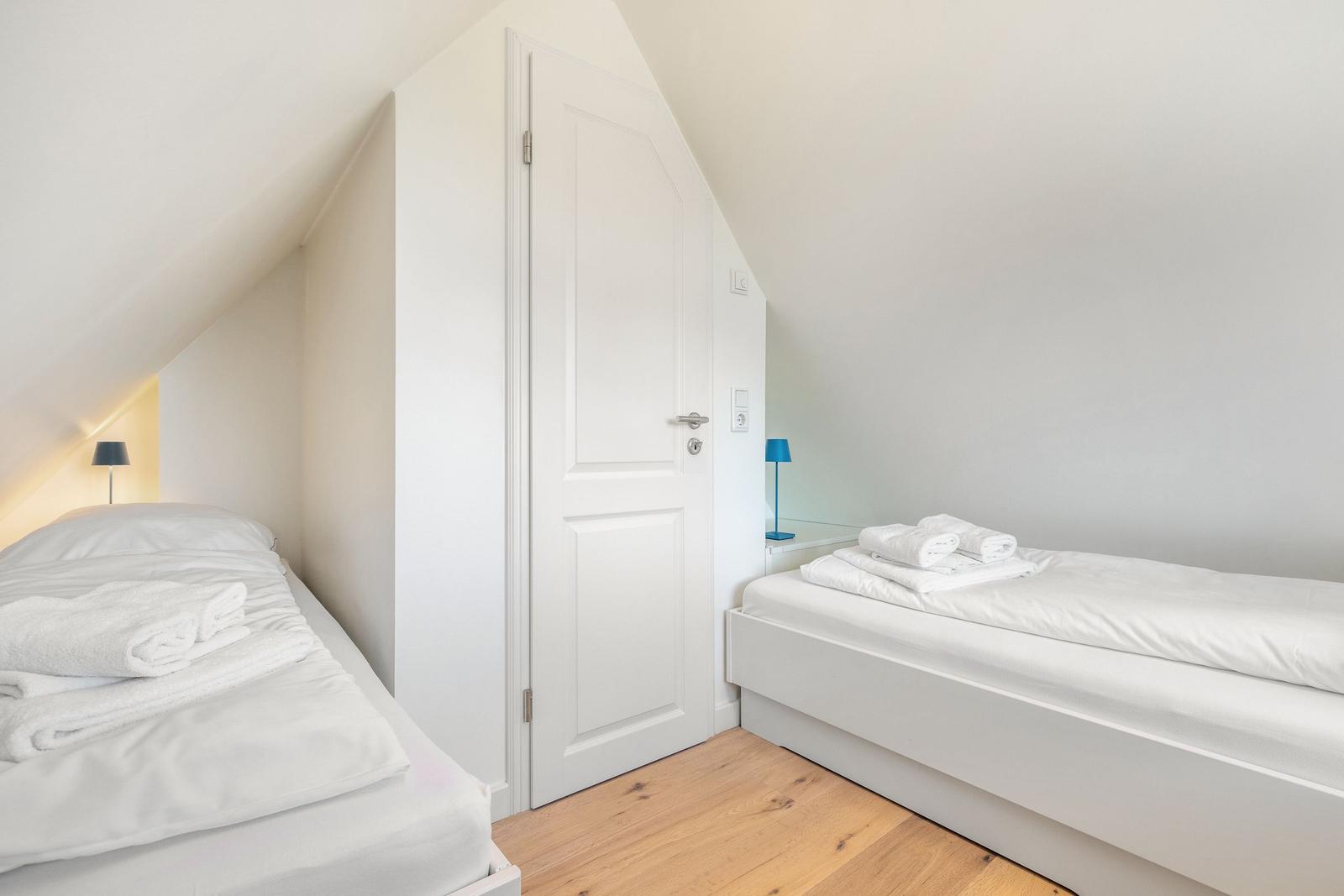 Zwei Betten mit weißen Bettwäsche und Handtüchern in einem Dachzimmer mit Holzboden und weißer Tür.