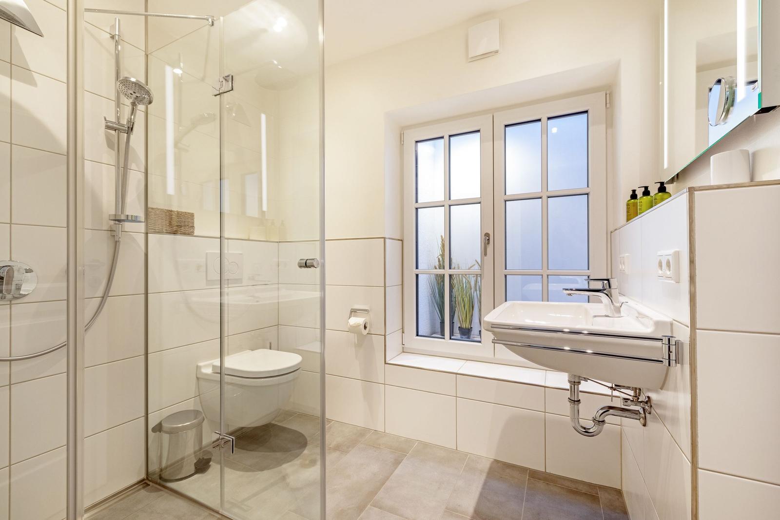Badezimmer mit Dusche, Toilette und Waschbecken. Fenster mit Pflanzen.
