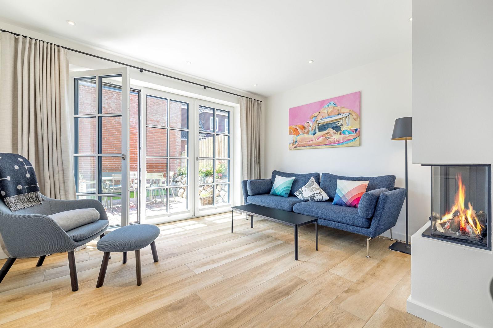 Wohnzimmer mit Sofa, Sessel, Kamin und Glasfront zur Terrasse.