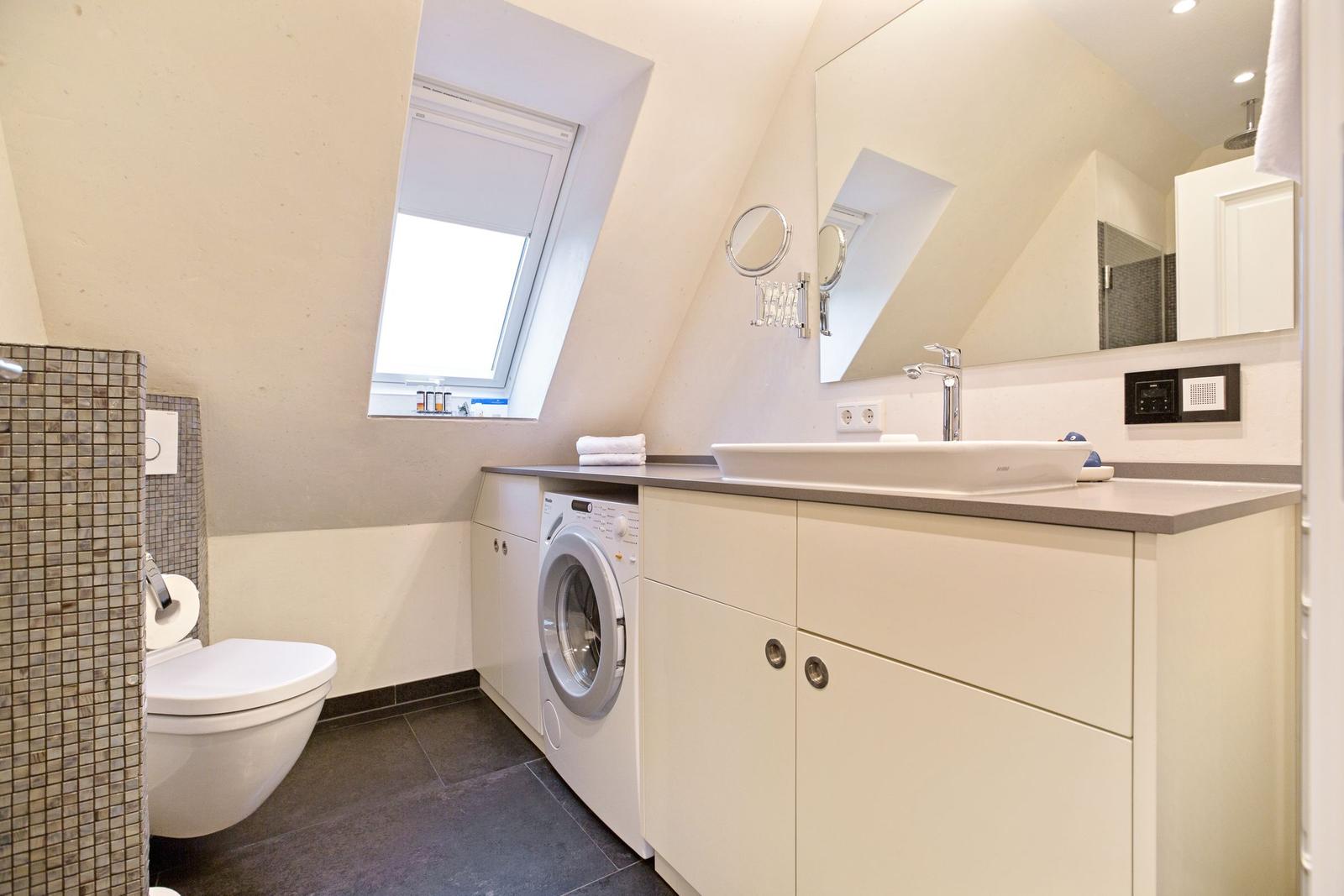 Badezimmer mit Waschmaschine, Waschtisch und Duschbereich unter Dachschrägen.