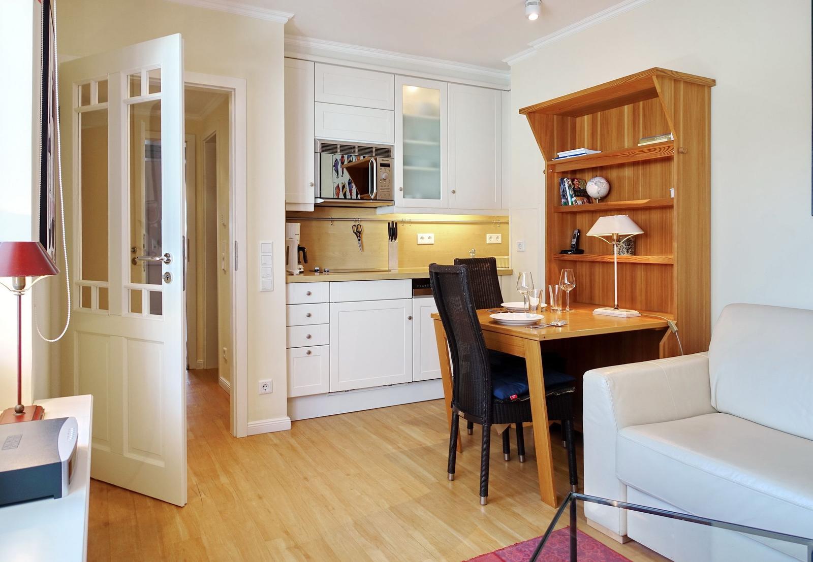 Apartment with kitchen, dining table, and sofa. Wooden floor and white furniture.