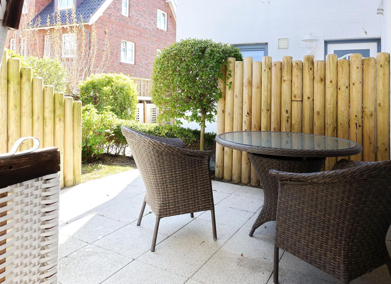 Terrace with round table and chairs, wooden fence, and green hedge in background.
