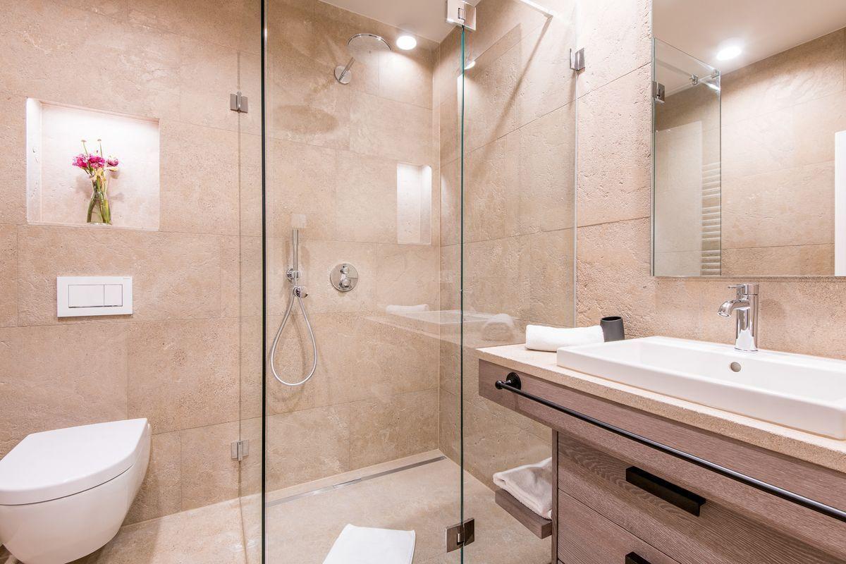 Bad mit Dusche, Waschbecken und Toilette. Glaswand, Holzschrank und Wandnische mit Blumen.