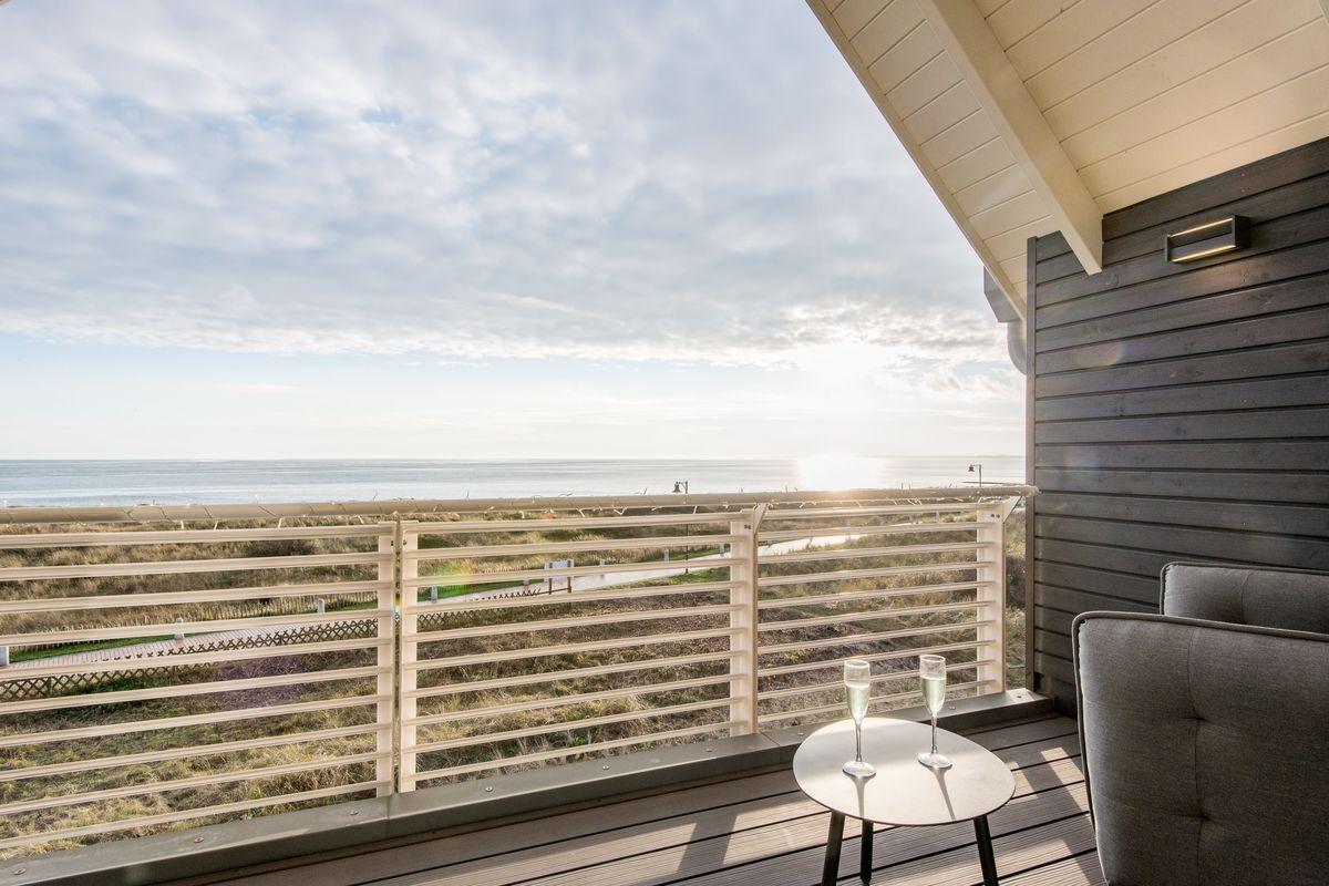 Terrasse mit Meerblick, Tisch und zwei Gläser. Holzgeländer und graue Sitzgelegenheit.