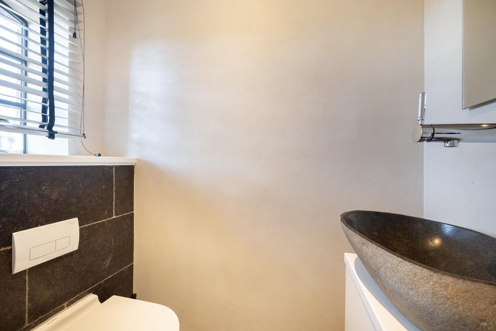 Bad mit Steinwaschbecken, Toilette und Fenster mit Jalousie.