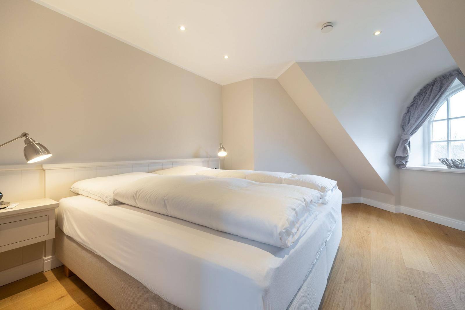 Schlafzimmer mit Doppelbett, Holzboden und Dachschrägen. Fenster mit grauem Vorhang.