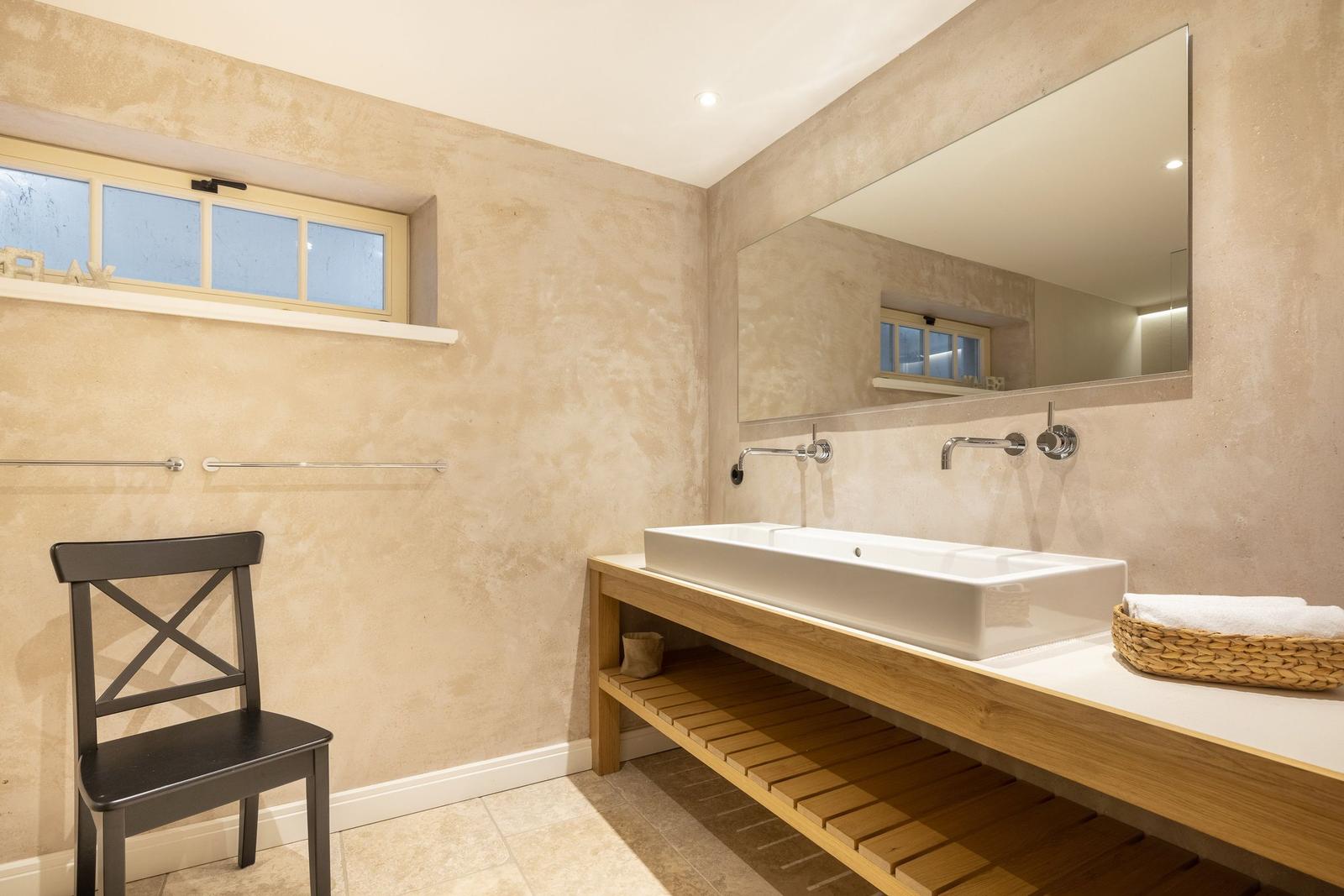 Badezimmer mit Waschtisch, Spiegel und Fenster. Holzregal und schwarze Stuhl.