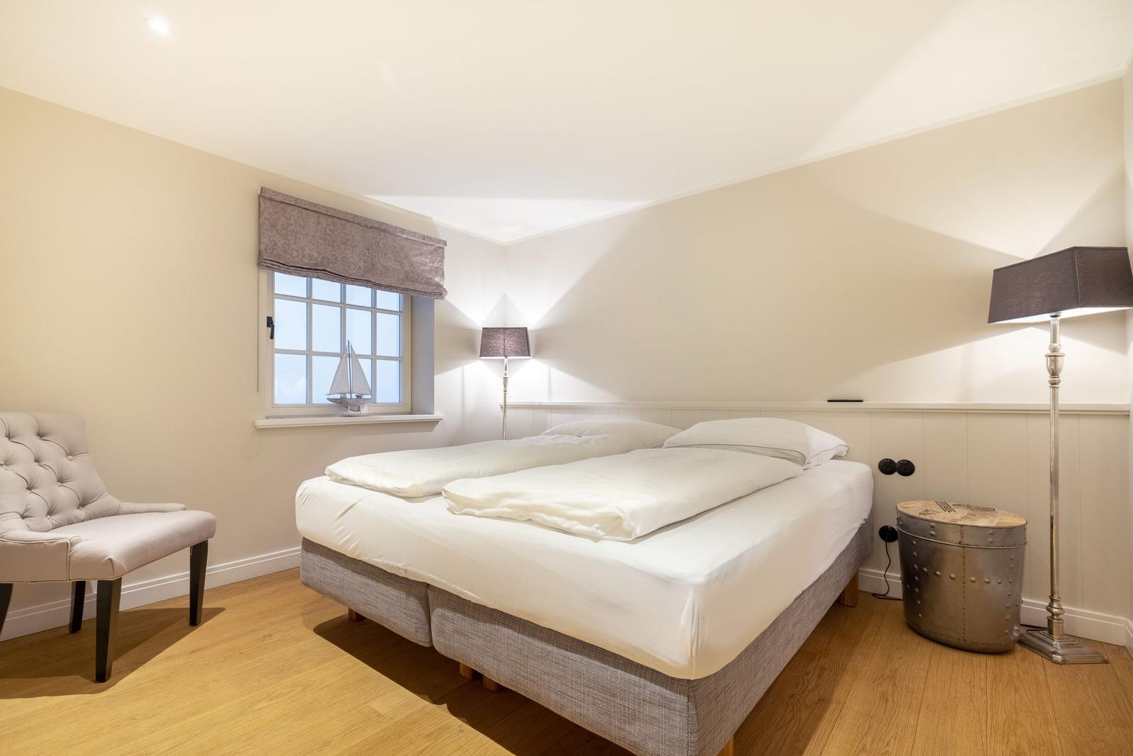 Schlafzimmer mit Bett, Stuhl und Fenster. Holzboden und Stehlampe.