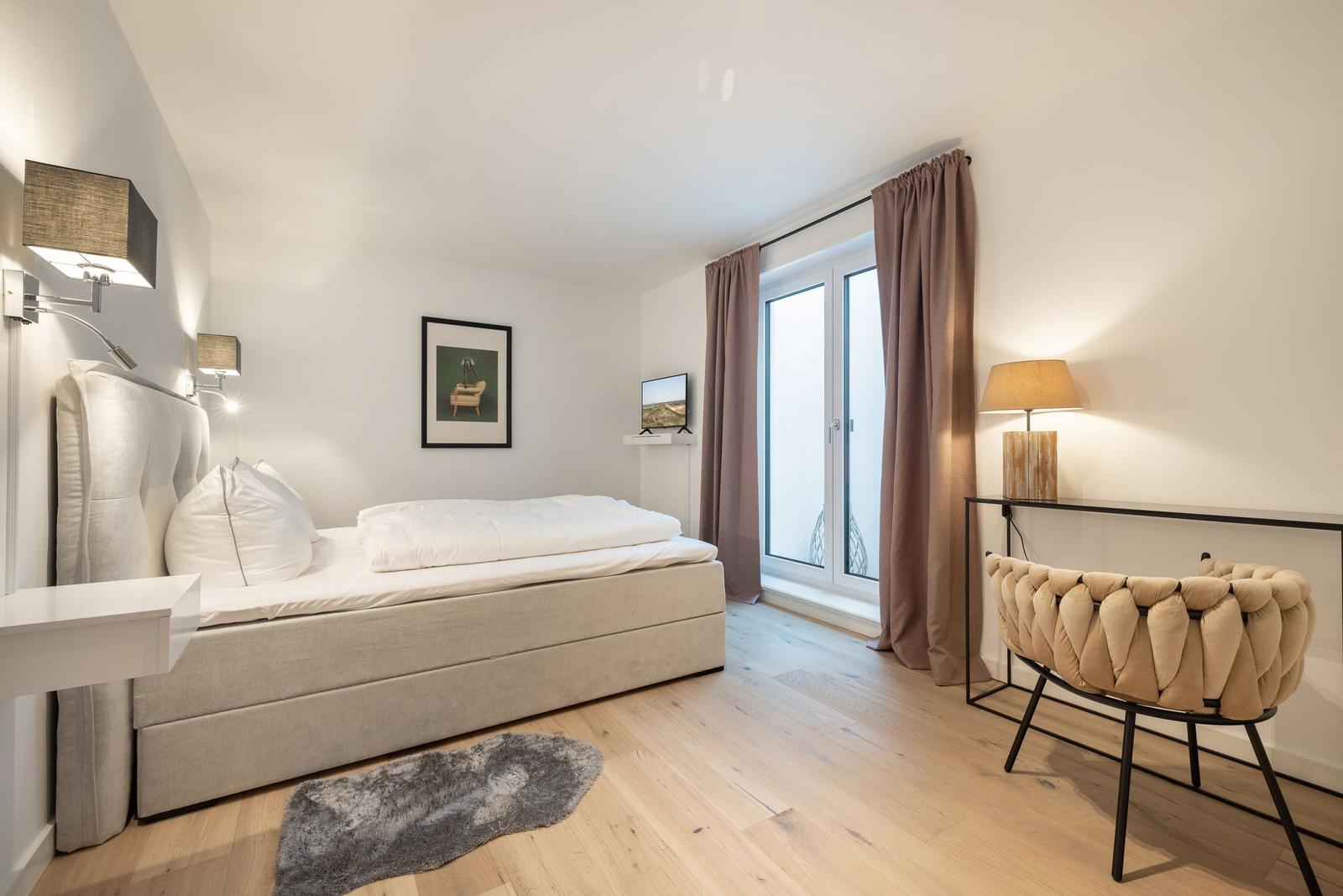 Schlafzimmer mit Bett, Schreibtisch und TV. Holzboden und große Fenster.