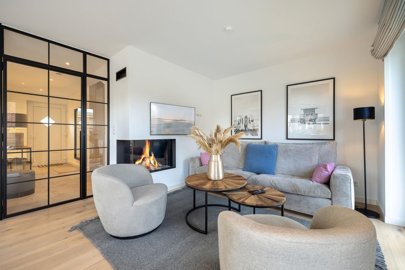 Wohnzimmer mit Kamin, Sofa und zwei Sesseln. Glaswand trennt den Raum.