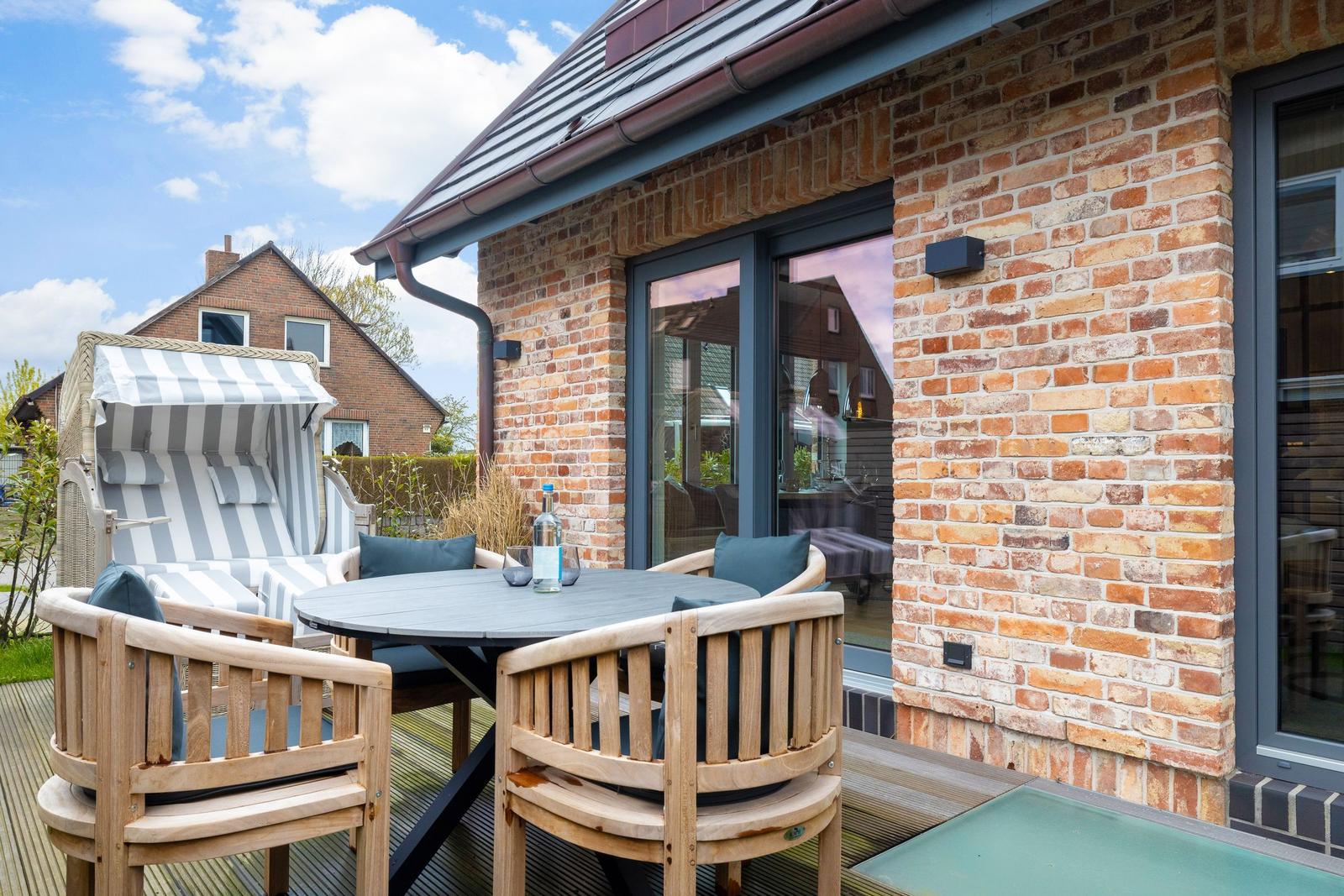 Terrasse mit Holztisch, Stühlen und Strandkorb. Backsteinhaus mit Glasfront.