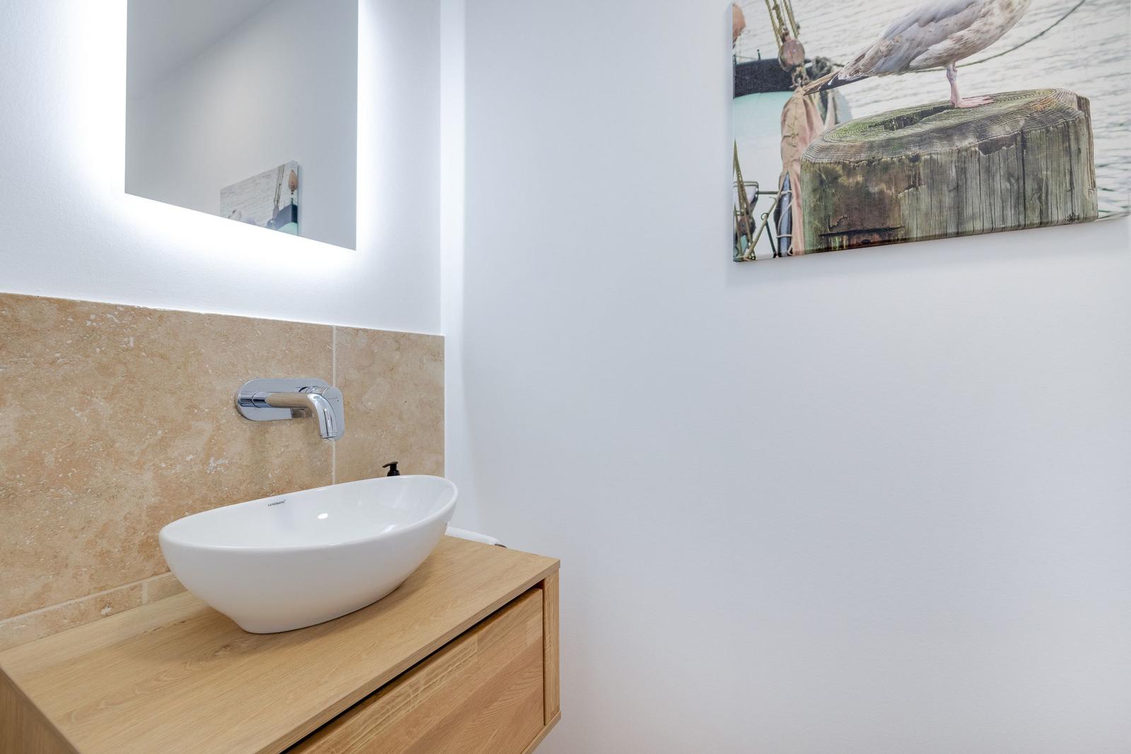 Waschbecken auf Holzunterschrank mit beleuchtetem Spiegel und Wandbild.
