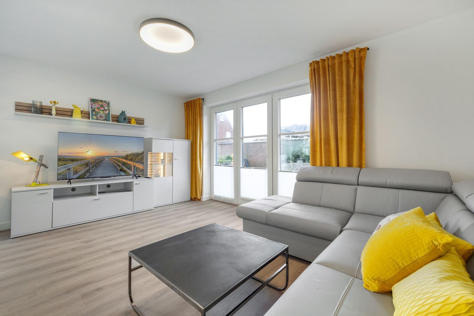 Wohnzimmer mit grauem Sofa, gelben Kissen und Fernseher.