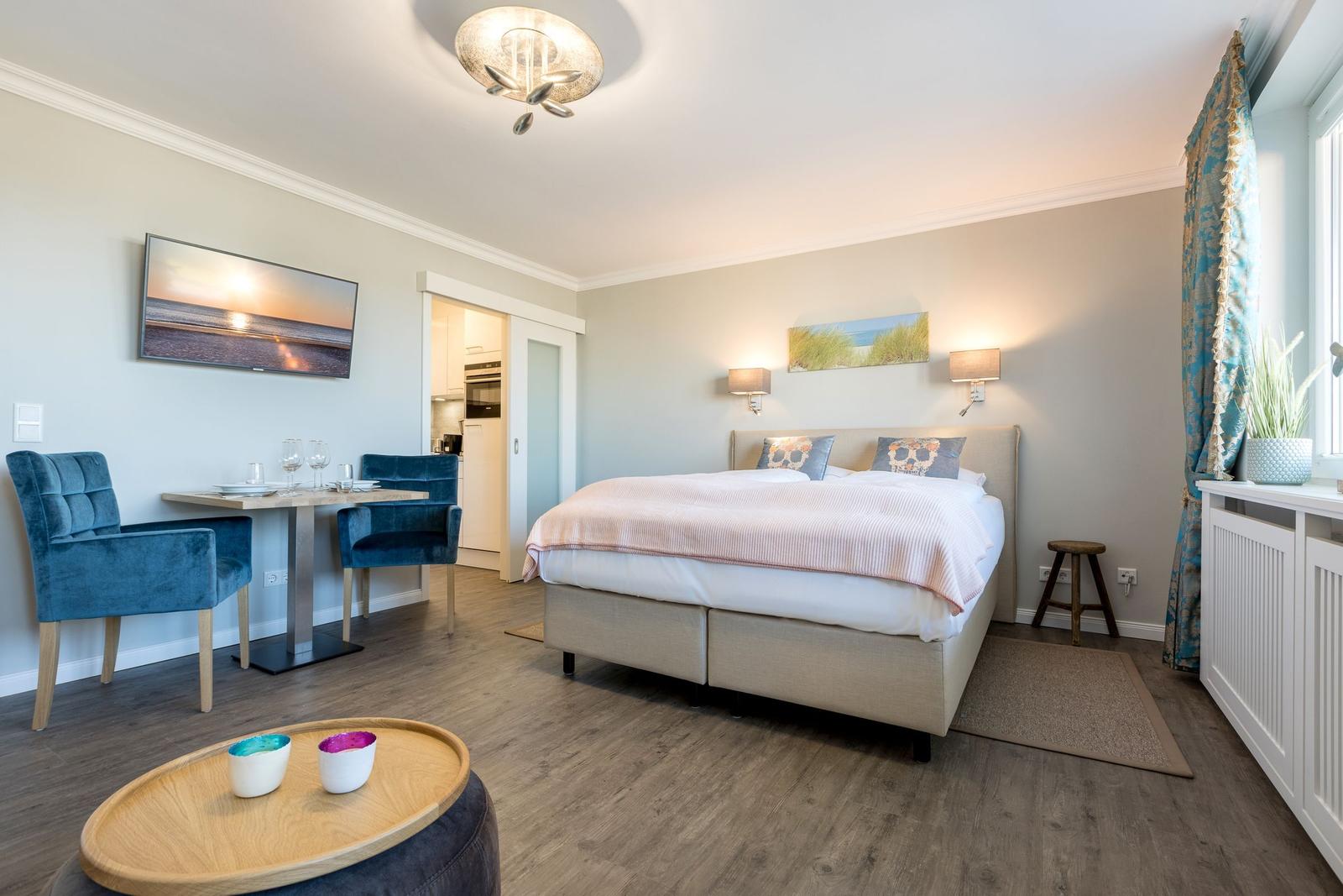Schlafzimmer mit Bett, Tisch, Stühlen und TV. Blick in die Küche.