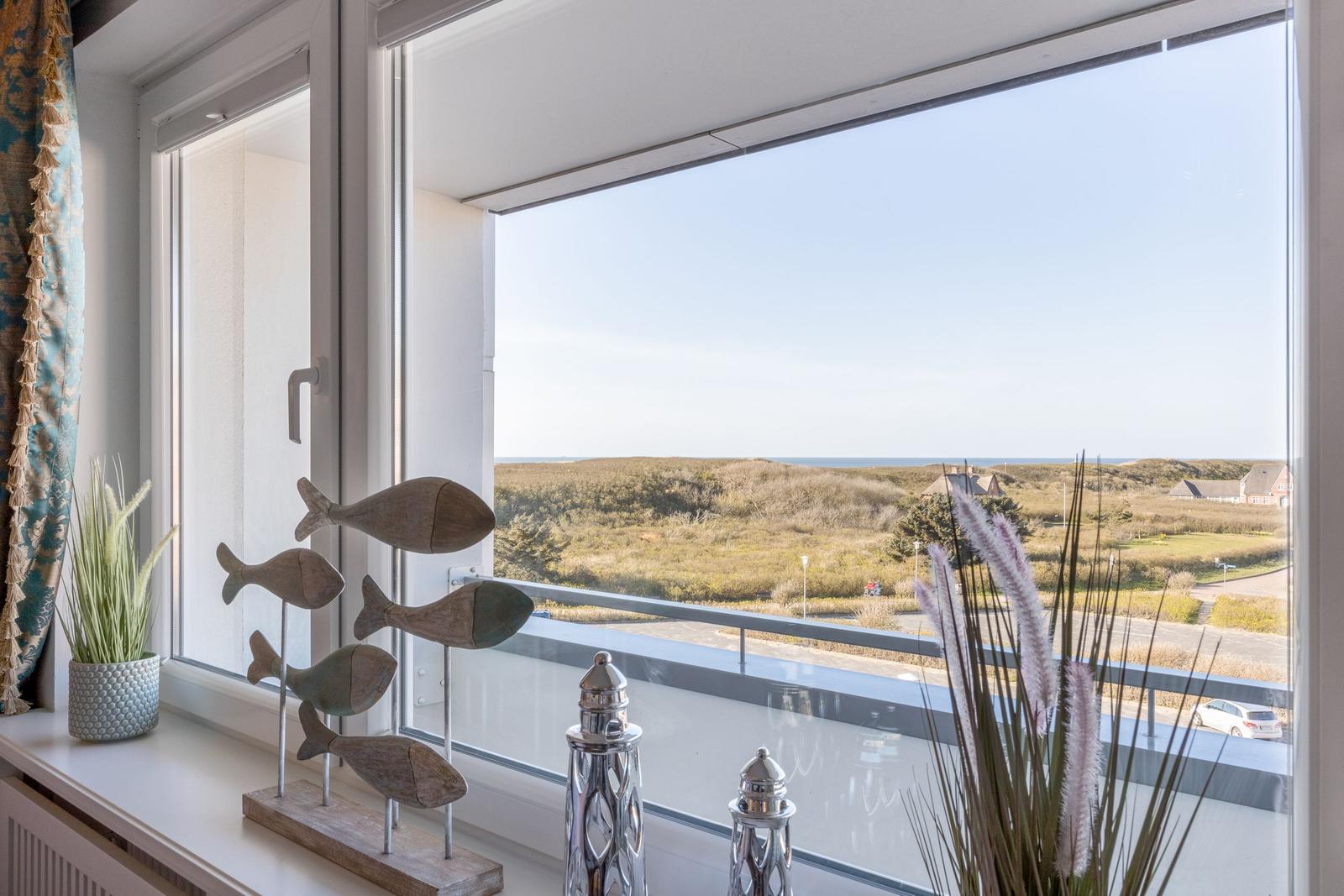 Blick auf Dünen und Meer durch große Fenster mit dekorativen Fischen und Pflanzen.