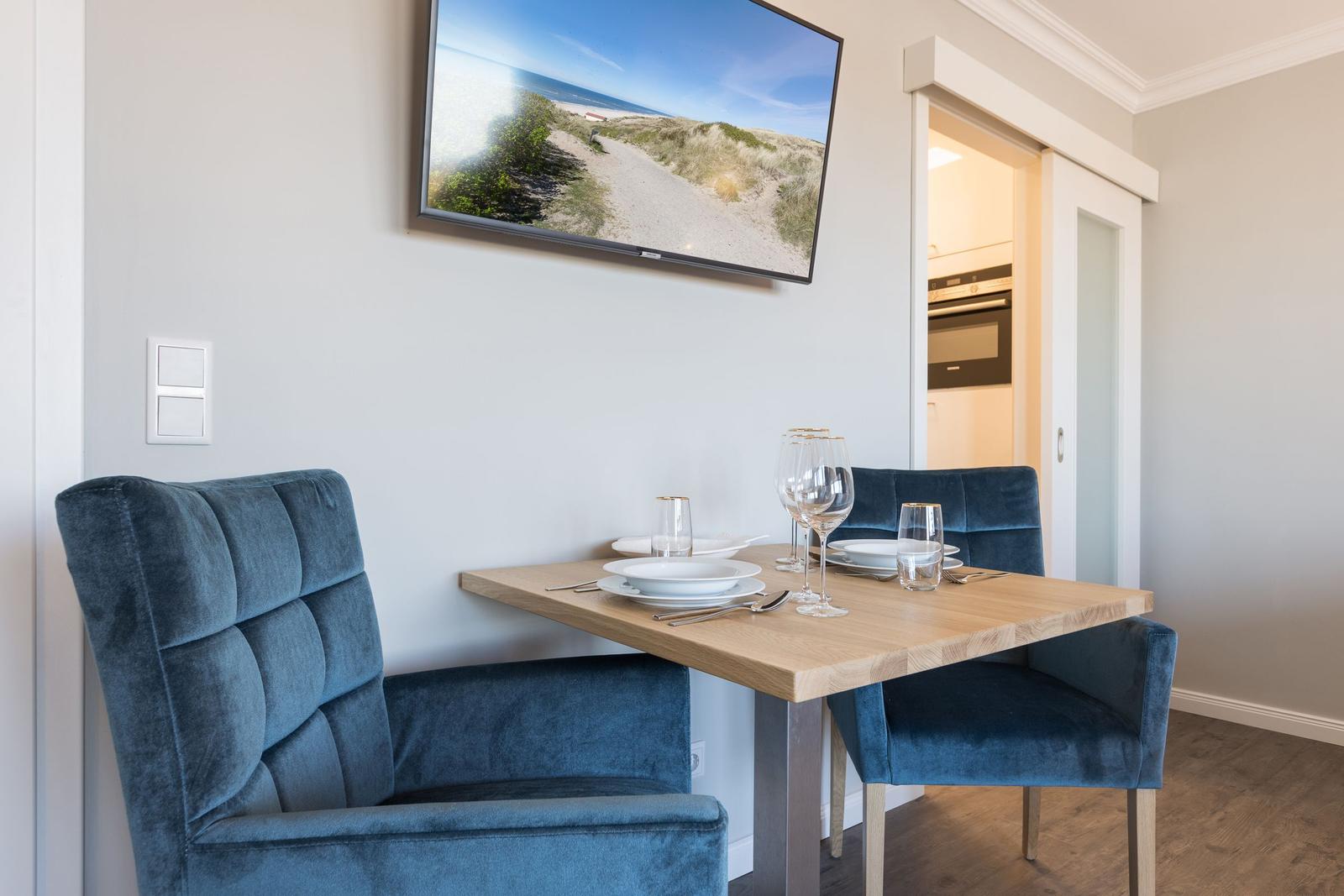 Essbereich mit Holztisch, blauen Stühlen und TV an der Wand.