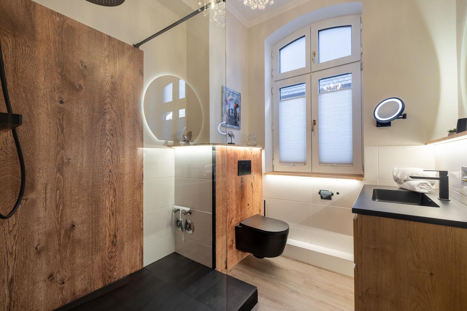 Badezimmer mit Dusche, Toilette und Waschbecken. Holzwände und großes Fenster.