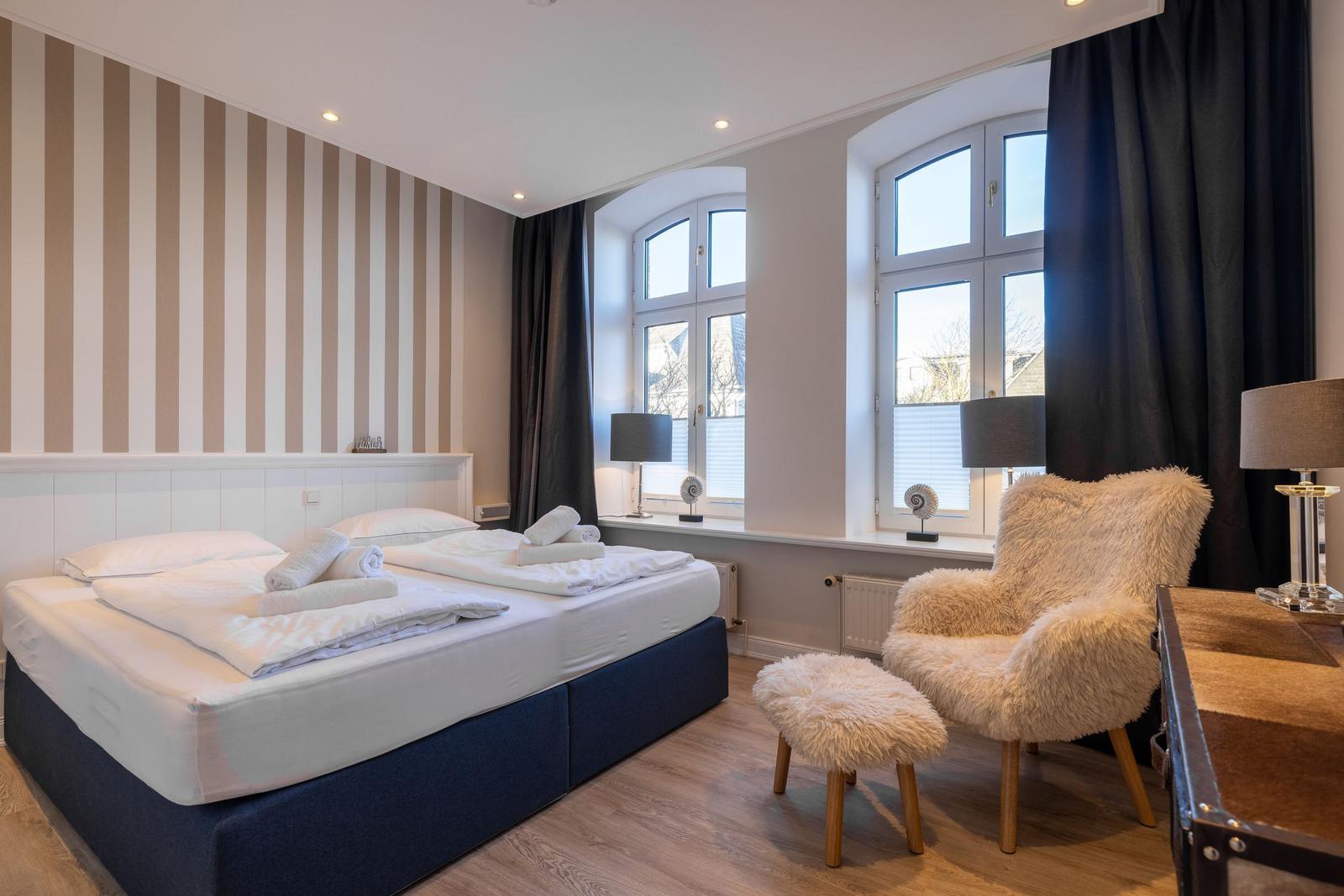 Schlafzimmer mit Bett, Fellstuhl und großen Fenstern.