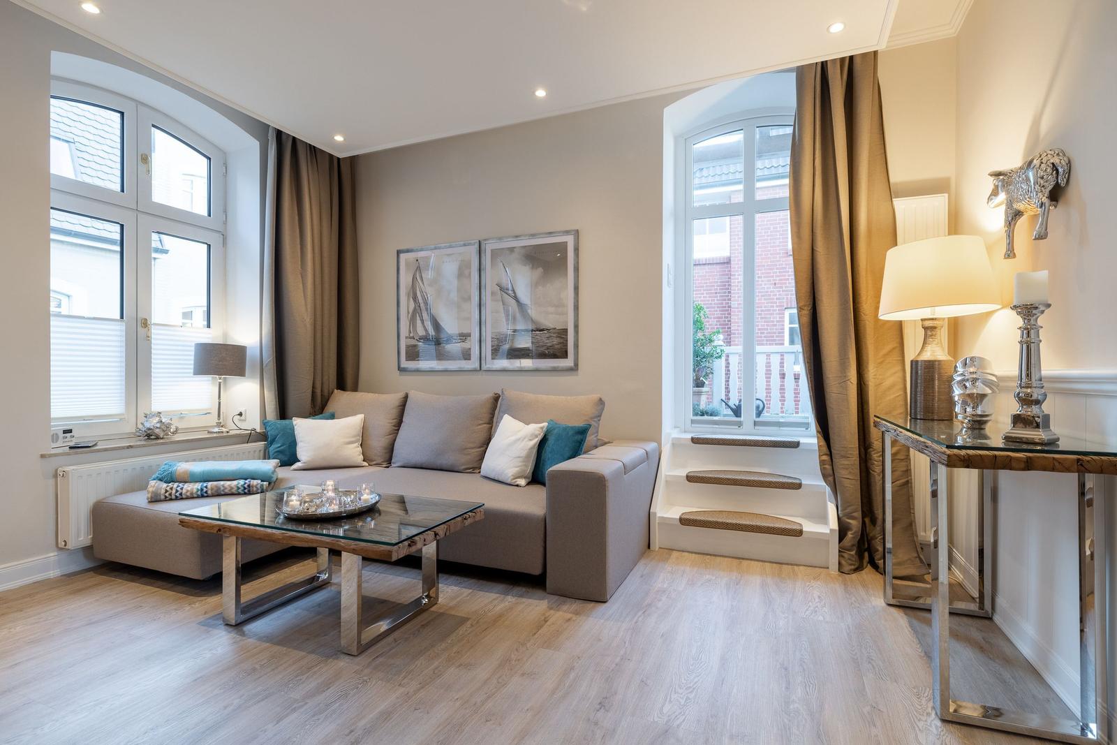 Wohnzimmer mit Sofa, Glas-Tisch und zwei großen Fenstern mit Blick auf Außenbereich.