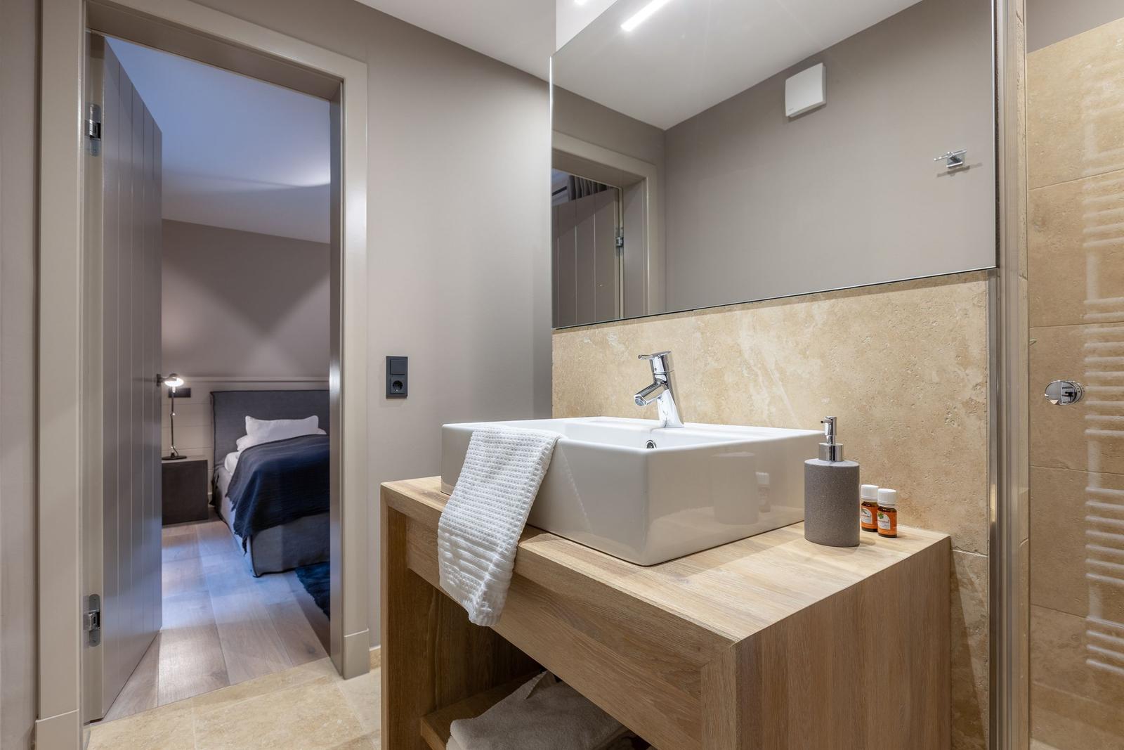 Badezimmer mit Waschbecken, Spiegel und Dusche. Schlafzimmer im Hintergrund sichtbar.