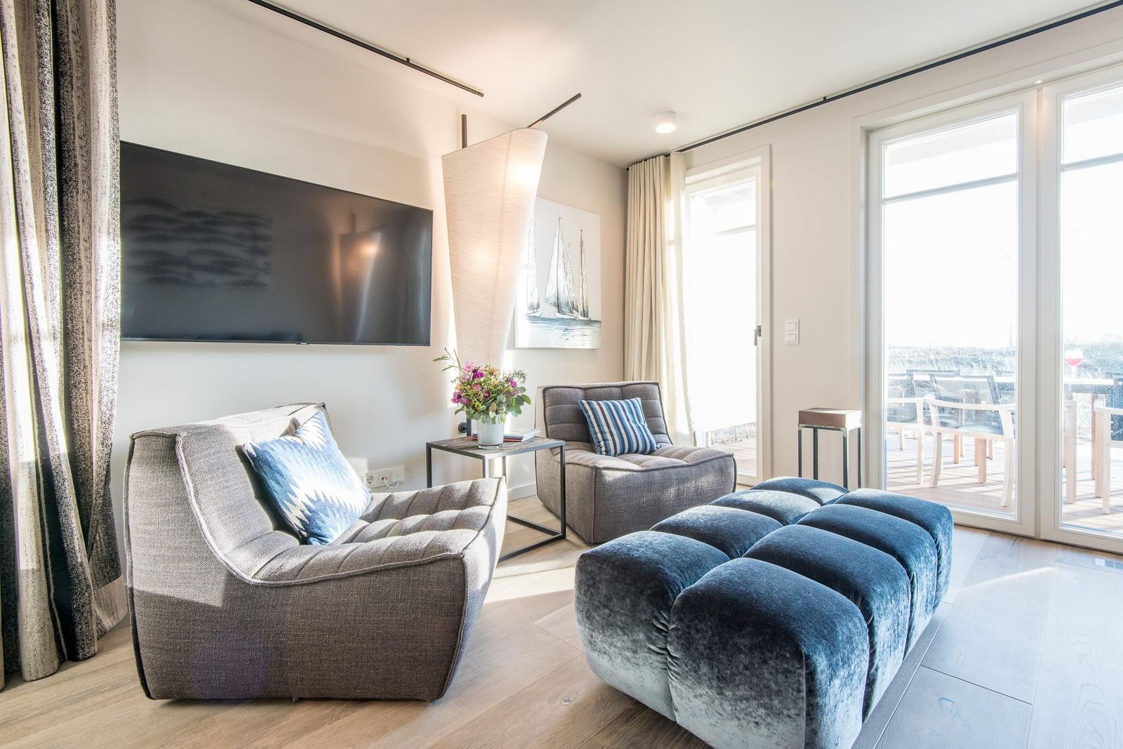 Wohnzimmer mit Sofa, Sessel und blauem Sitzkissen. Große Fenster und Fernseher an der Wand.