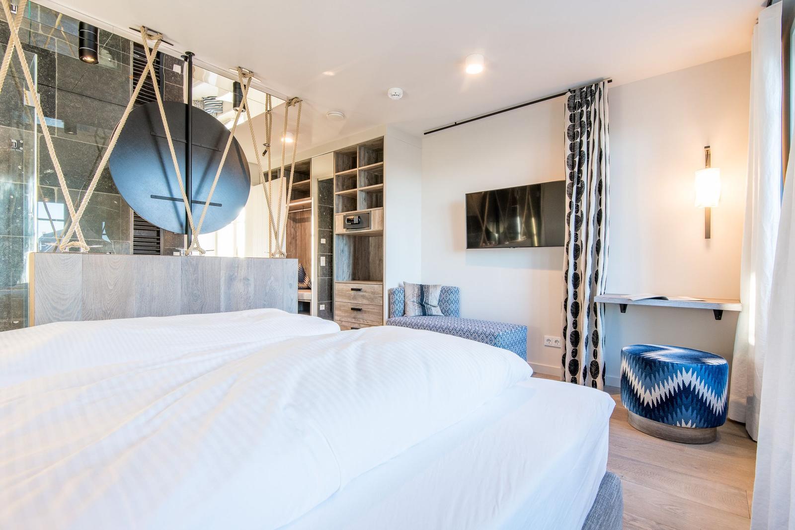 Schlafzimmer mit Bett, Kleiderschrank und TV. Blaue Sitzgelegenheit und dekorative Vorhänge.