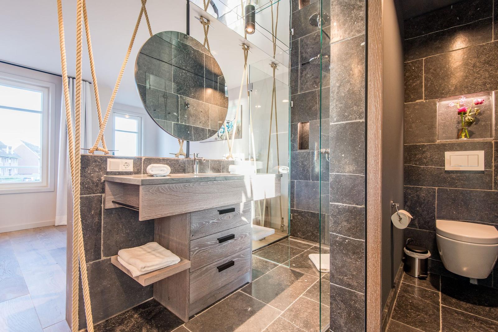 Badezimmer mit Dusche, Waschbecken und Toilette. Dunkle Fliesen und Holzmöbel.