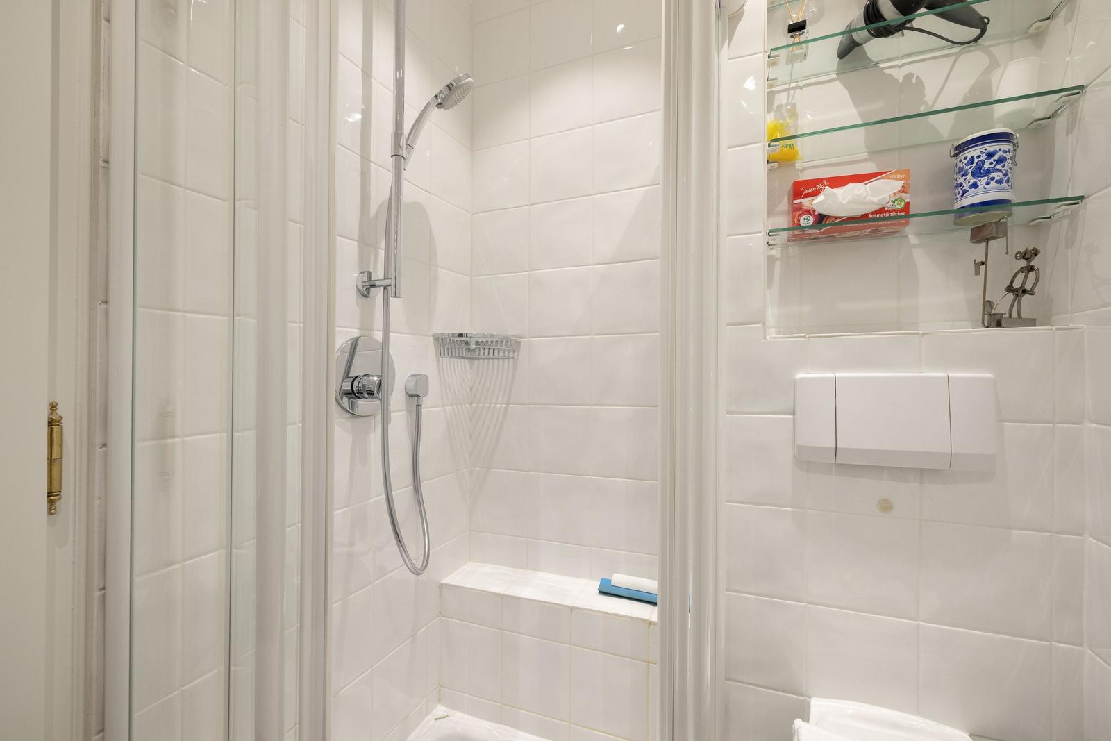 Shower with glass enclosure, showerhead, and shelf with tissue box and hair dryer.