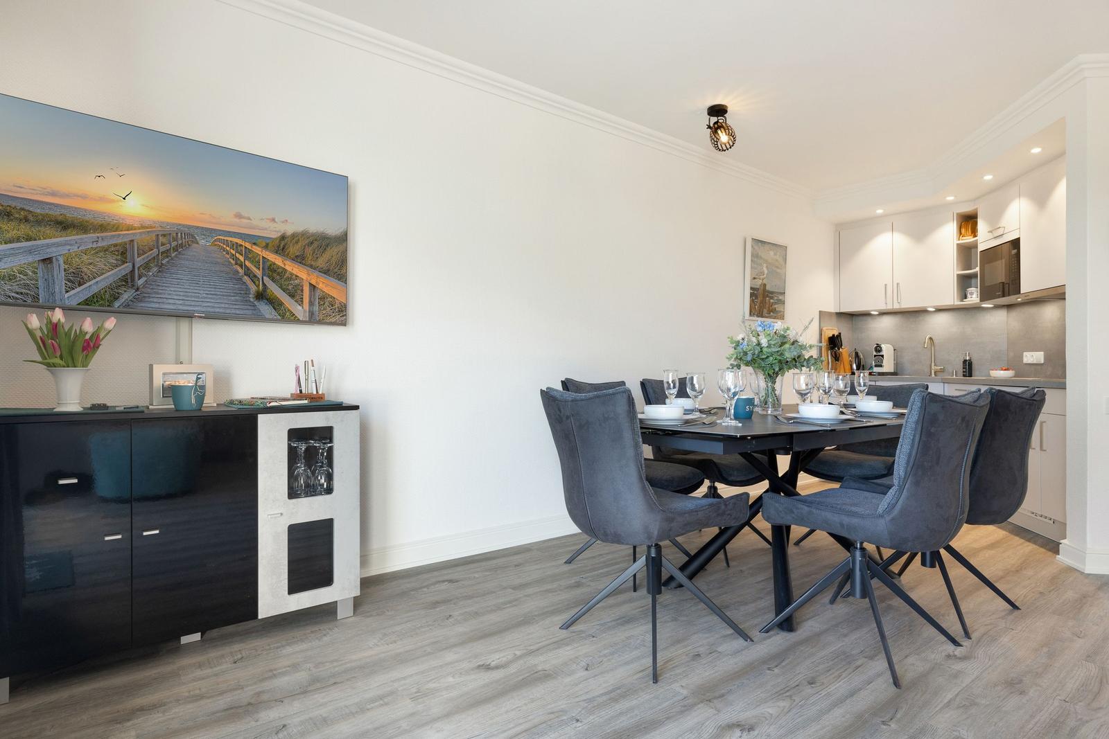 Dining area with table, chairs, and kitchen. Wall art shows beach.