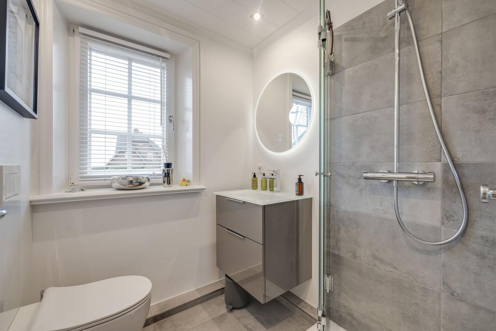 Badezimmer mit Dusche, Waschbecken und Toilette. Fenster mit Jalousie.