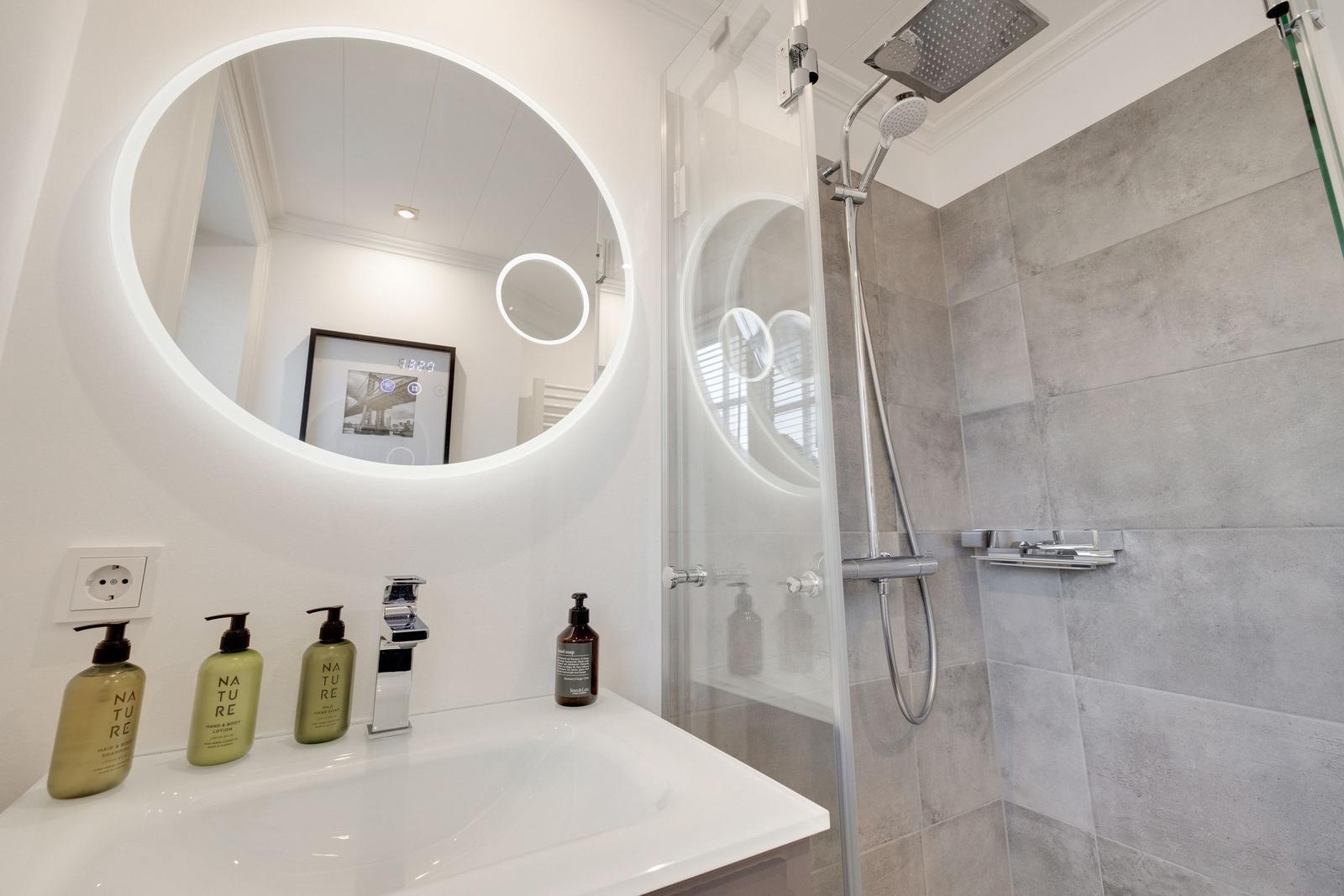 Badezimmer mit runden Spiegel, Dusche und Waschbecken mit Seifenflaschen.