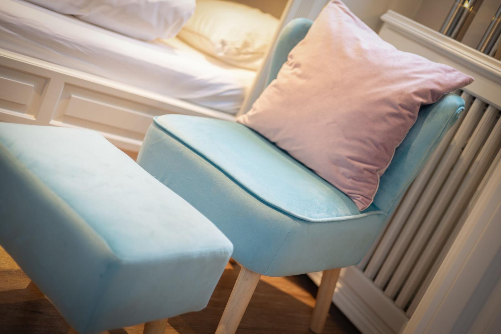 Ein hellblauer Sessel mit Kissen und Hocker neben dem Bett.