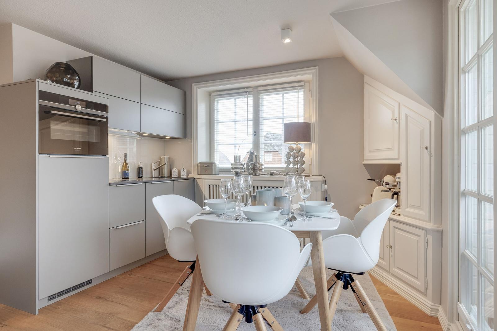 Küche mit Essbereich, weißer Tisch und Stühle, Fenster mit Jalousien und moderner Einbauküche.