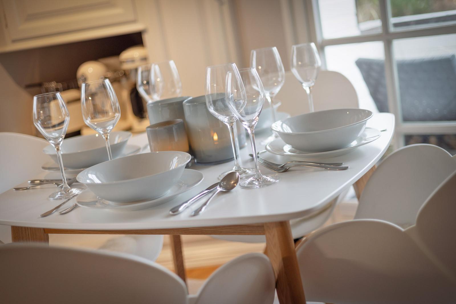 Tisch mit weißen Tellern, Gläsern und Besteck. Weiße Stühle und Fenster im Hintergrund.