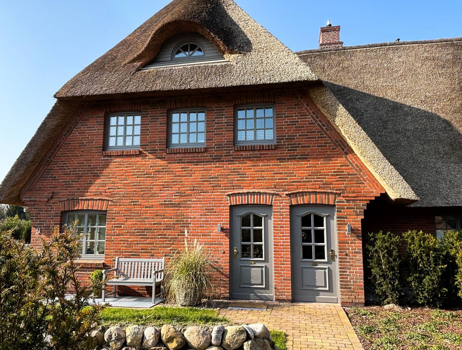 Rotes Backsteinhaus mit Strohdach, zwei Türen und Fenstern, Gartenbank und Beete.