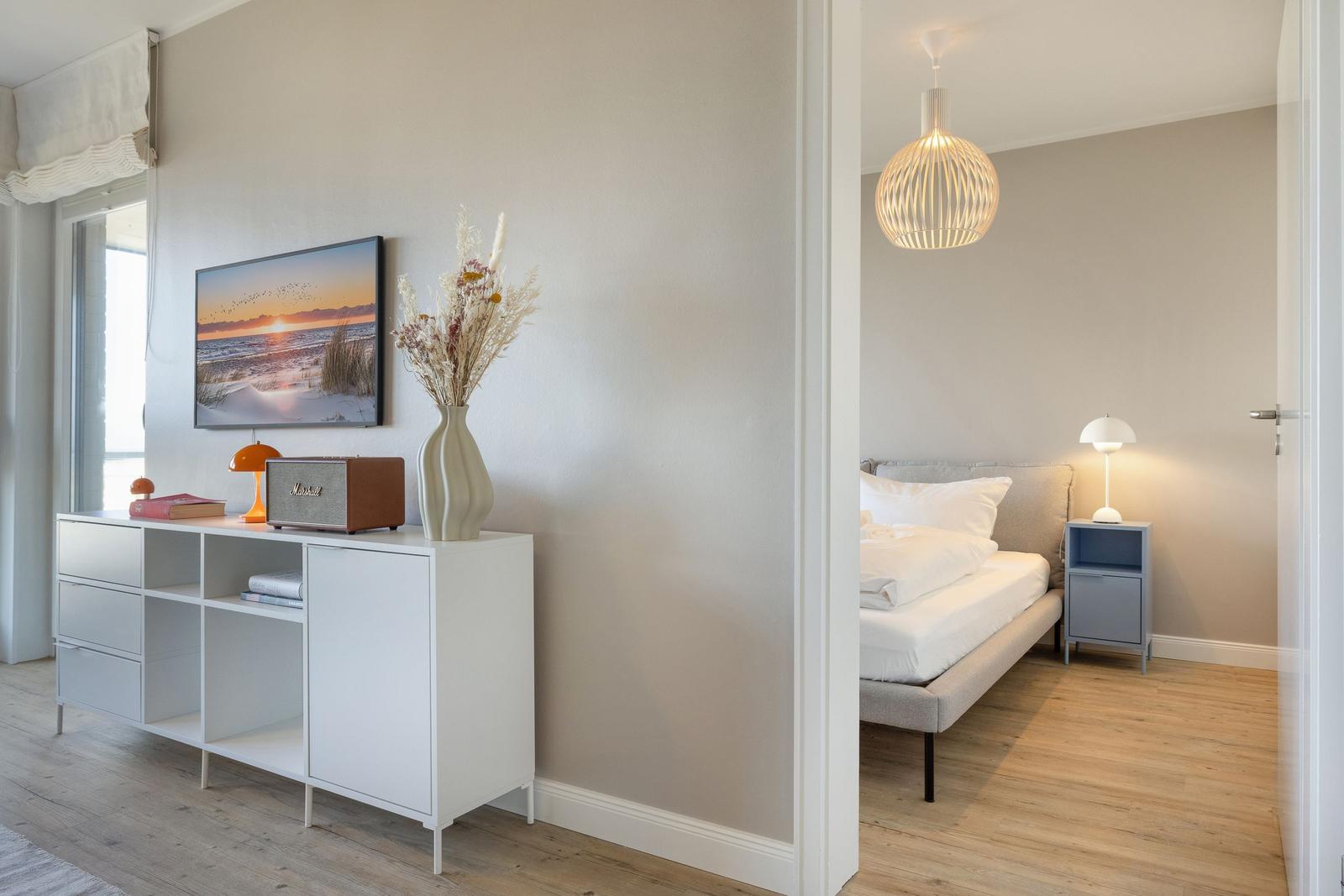 Schlafzimmer mit Bett, Nachttisch und Hängelampe. Nebenraum mit Sideboard, Bild und Vasen.
