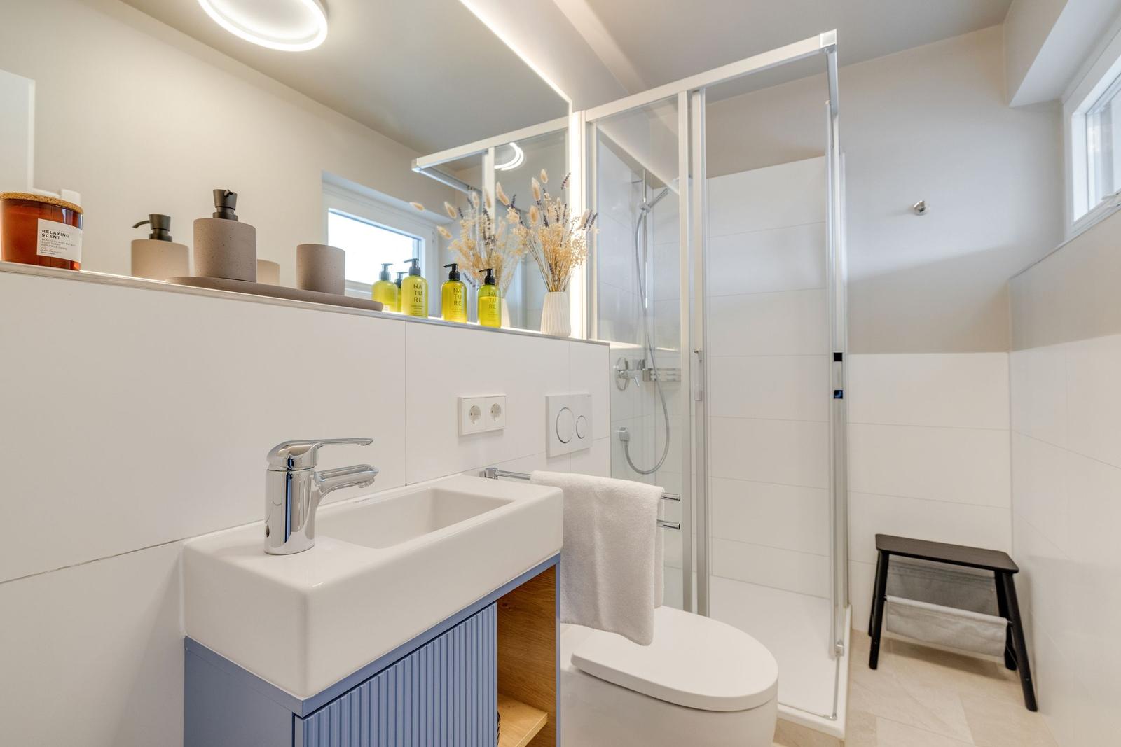 Badezimmer mit Dusche, Waschbecken und Toilette. Weiße Fliesen und moderne Einrichtung.