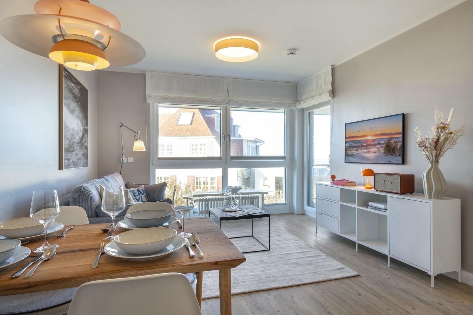 Wohn- und Essbereich mit Tisch, Sofa und TV. Große Fenster mit Blick auf Gebäude.
